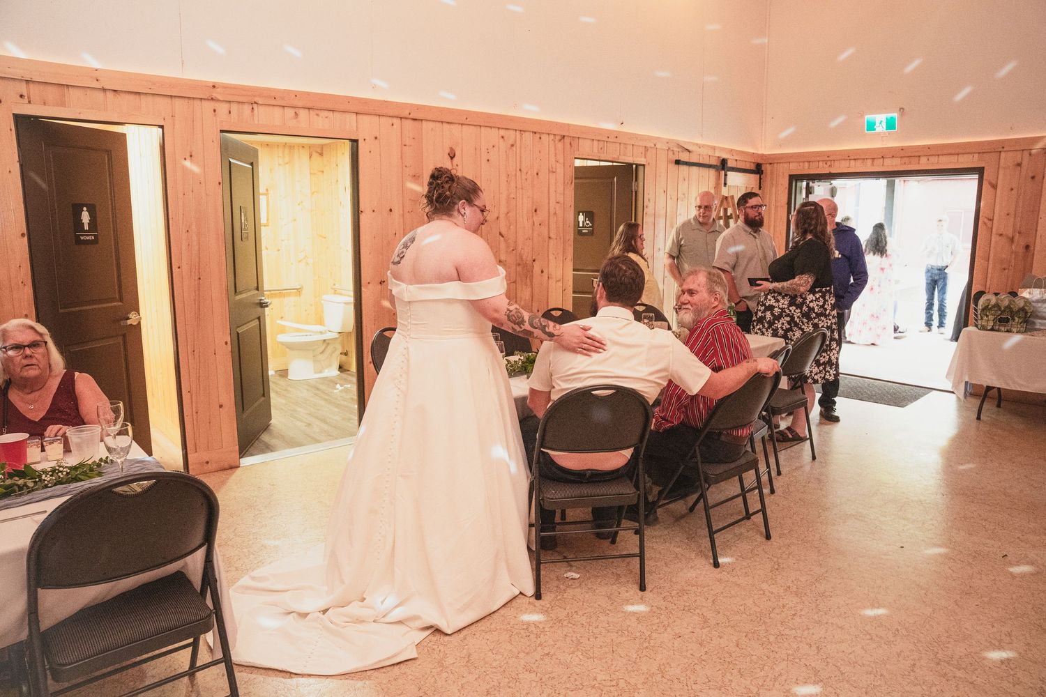 Bride in white gown greeting seated guests at a rustic indoor wedding reception.