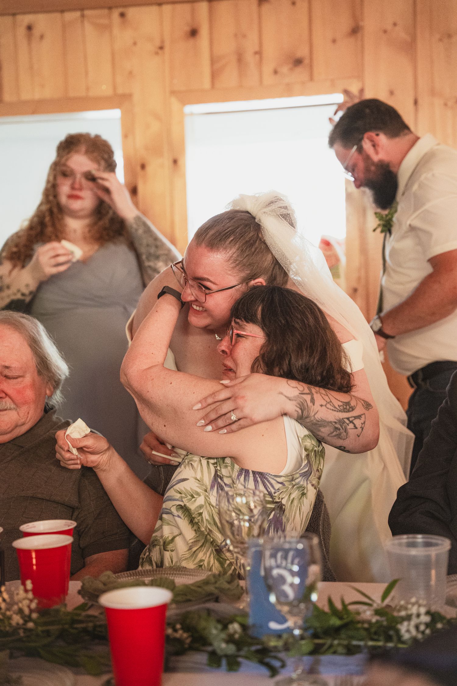 Bride in veil sharing an emotional hug with a guest during wedding reception in a wood-paneled venue.