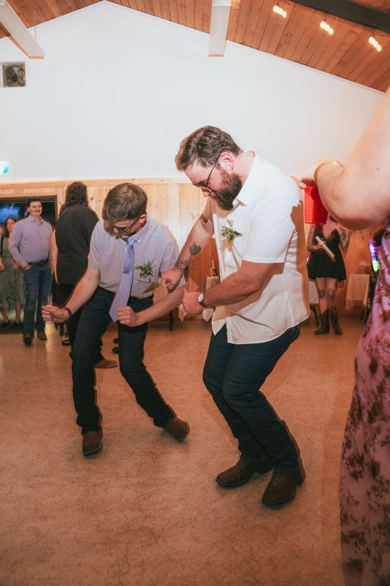Two men dancing energetically at a wedding reception in a rustic wood-paneled venue.
