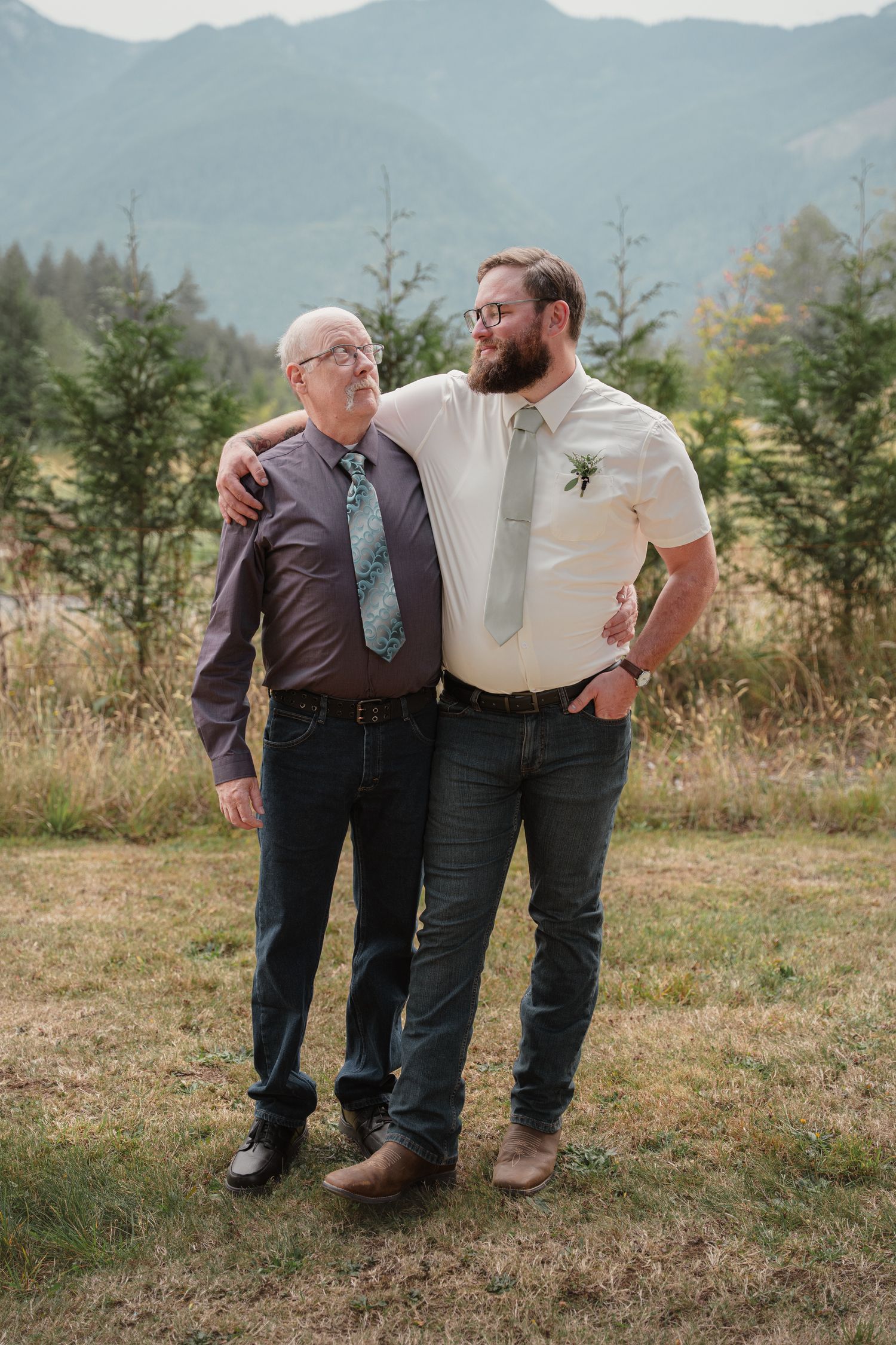 An older man and younger bearded groom share an embrace outdoors with mountain backdrop.