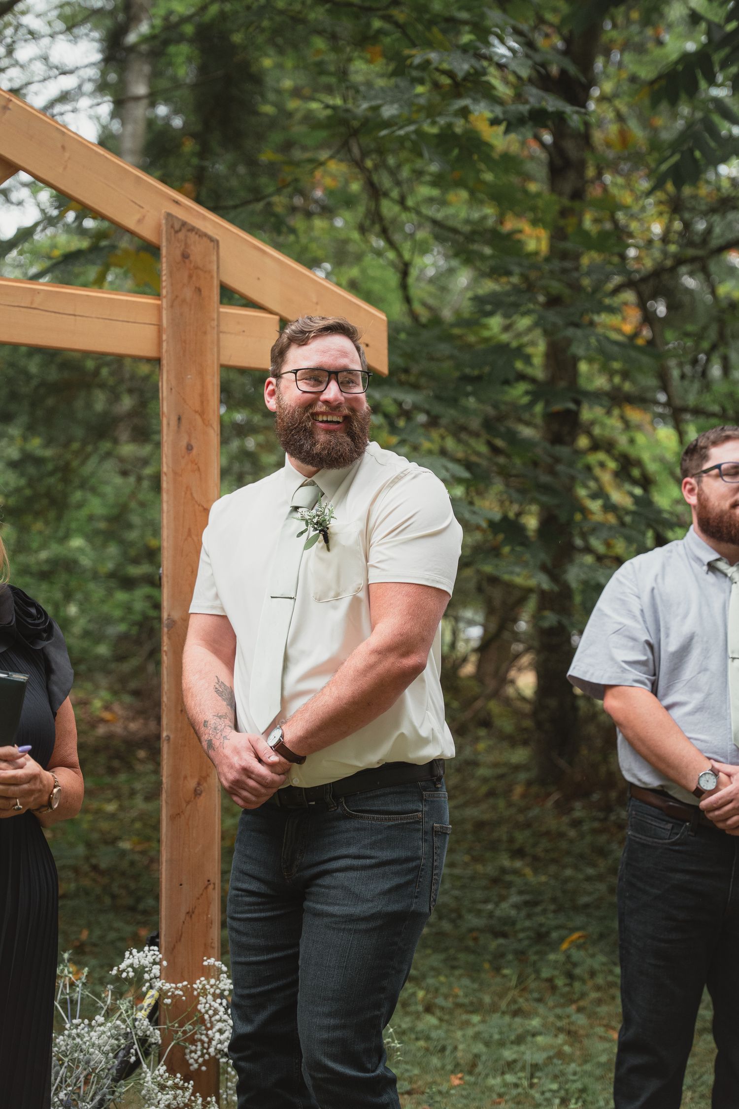 Bearded groom smiling joyfully at outdoor wedding ceremony beside wooden arch, wearing cream shirt with green floral tie.