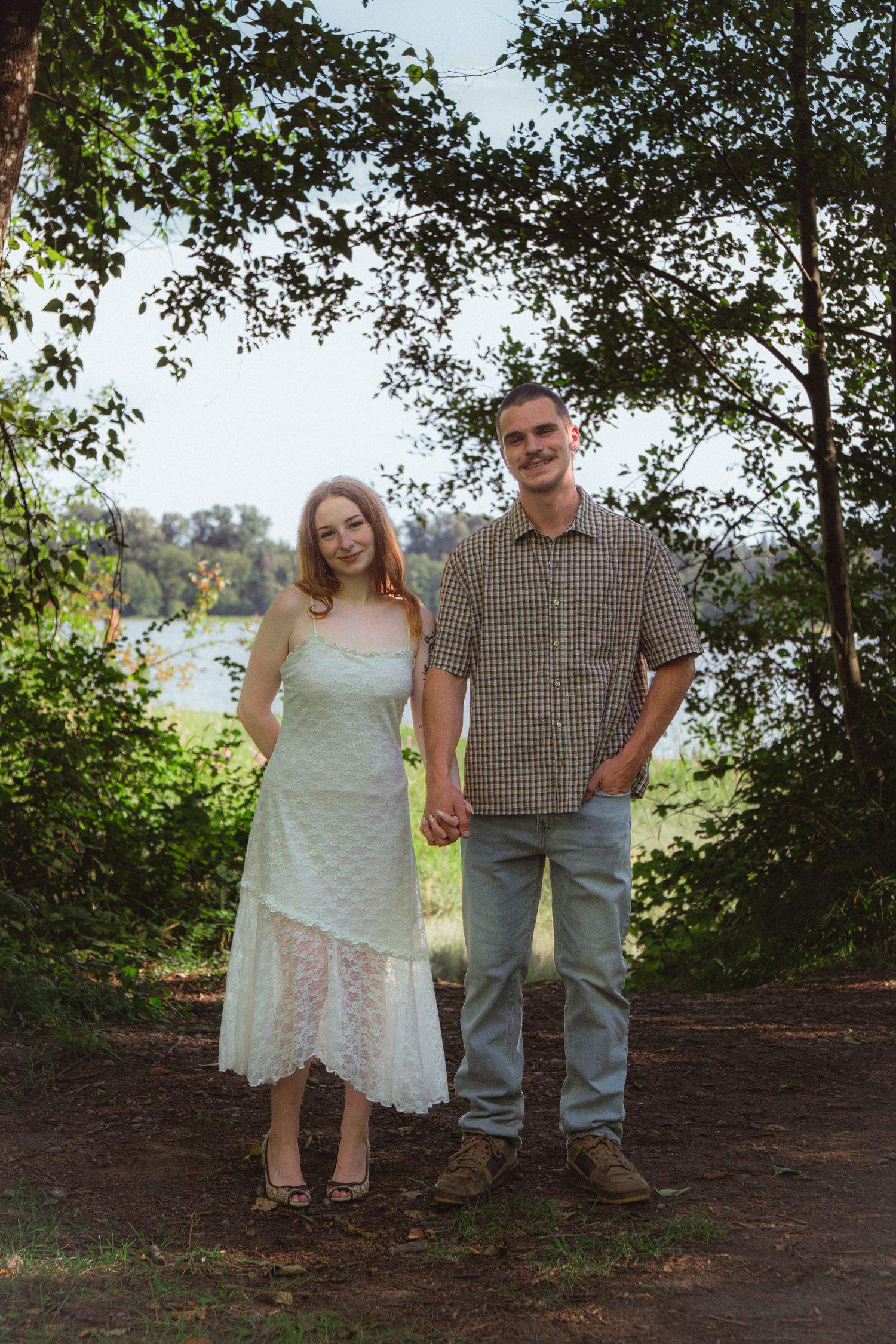 A couple holds hands on a wooded path near a river, woman in white dress, man in plaid shirt and jeans.