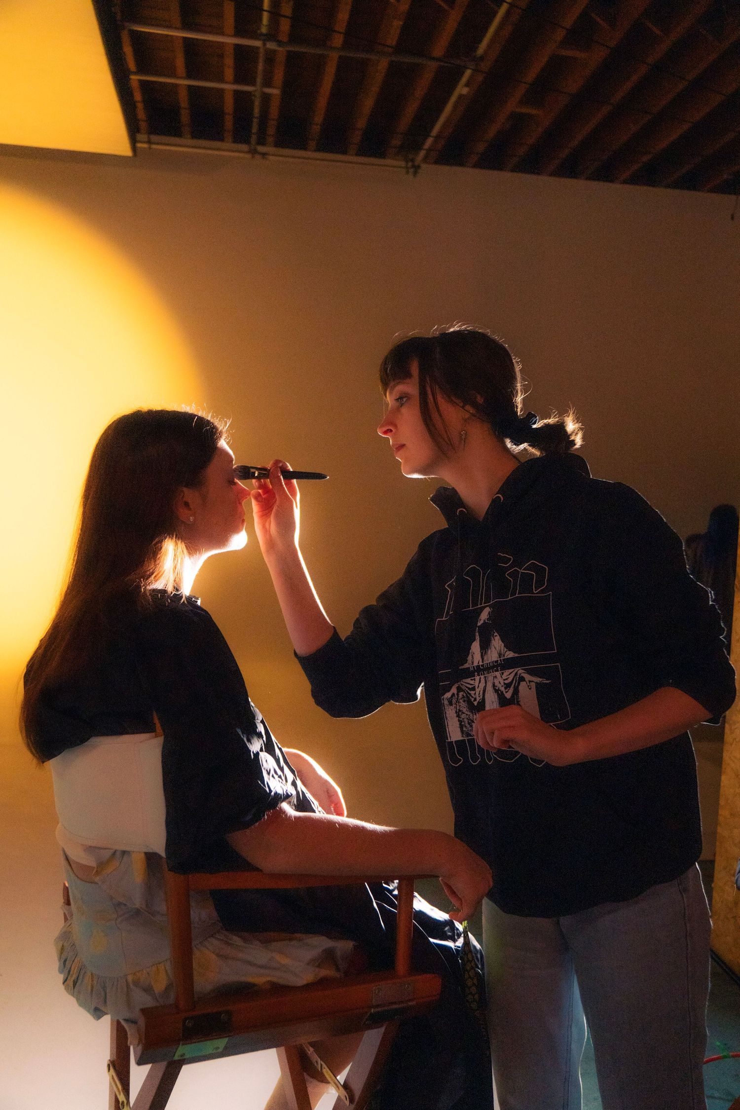 Makeup artist applying cosmetics to a seated actress on a warmly lit film set.