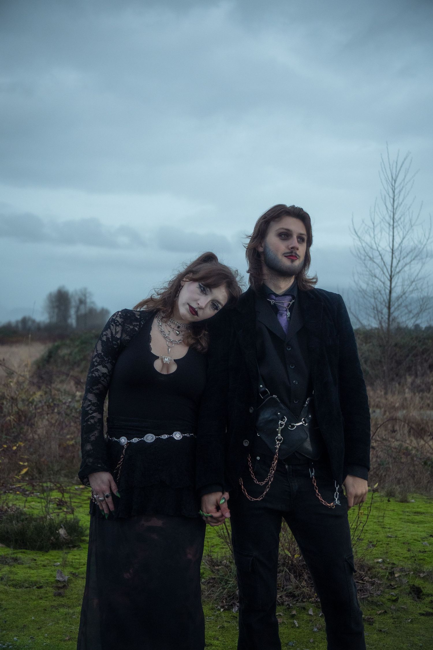 Gothic couple dressed in black standing in a moody overcast field landscape.