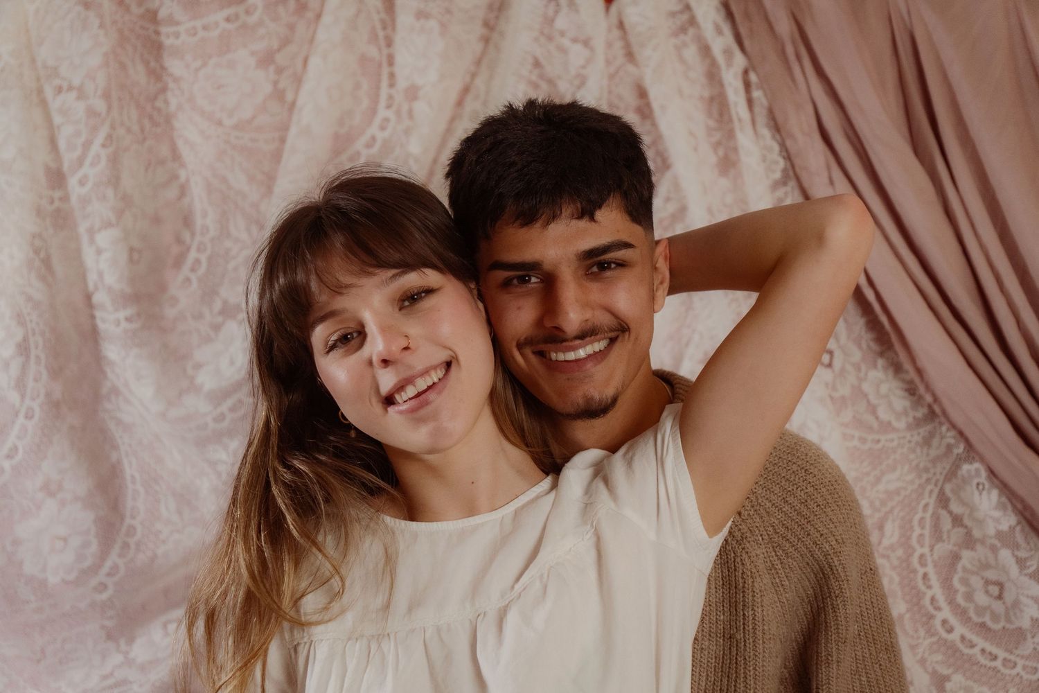 Smiling young couple posing together against a soft pink draped fabric backdrop in a romantic portrait.