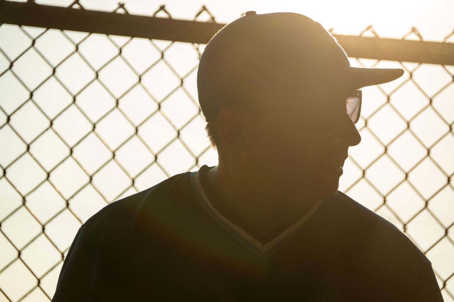 Silhouette of a baseball player wearing a cap standing against a chain-link fence in bright sunlight.