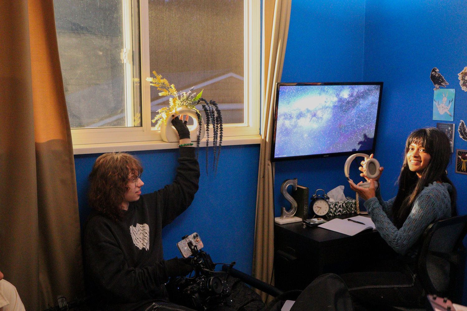 Two people on a film set hold circular props while sitting in a blue-walled room.