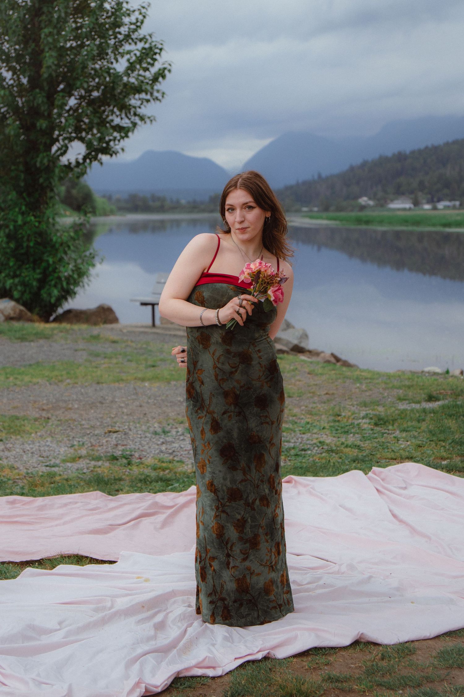 Woman in dark floral maxi dress with red top holds pink flowers by a mountain lake.