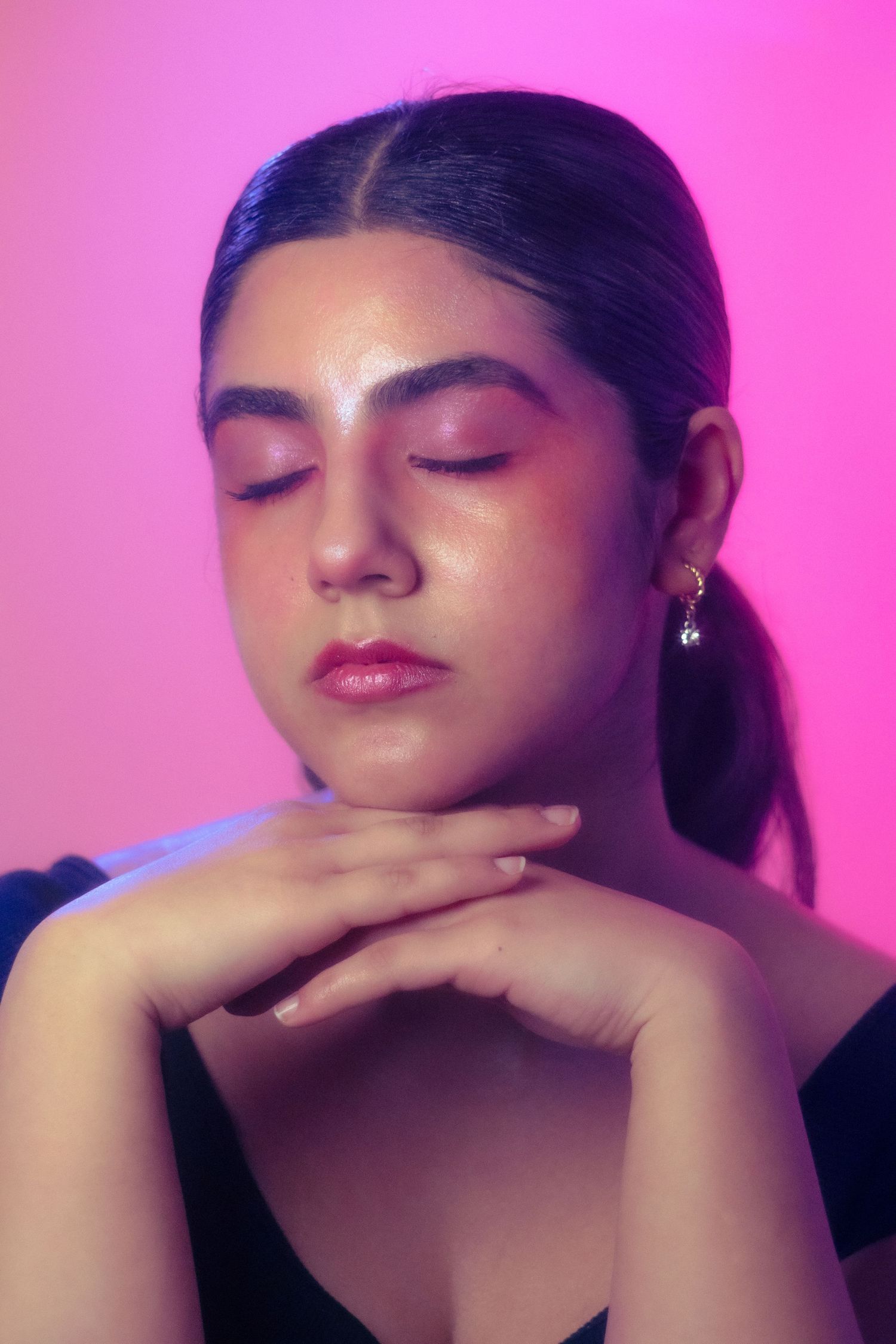 Close-up portrait of a young woman with pink eye makeup and closed eyes against a vibrant pink background.