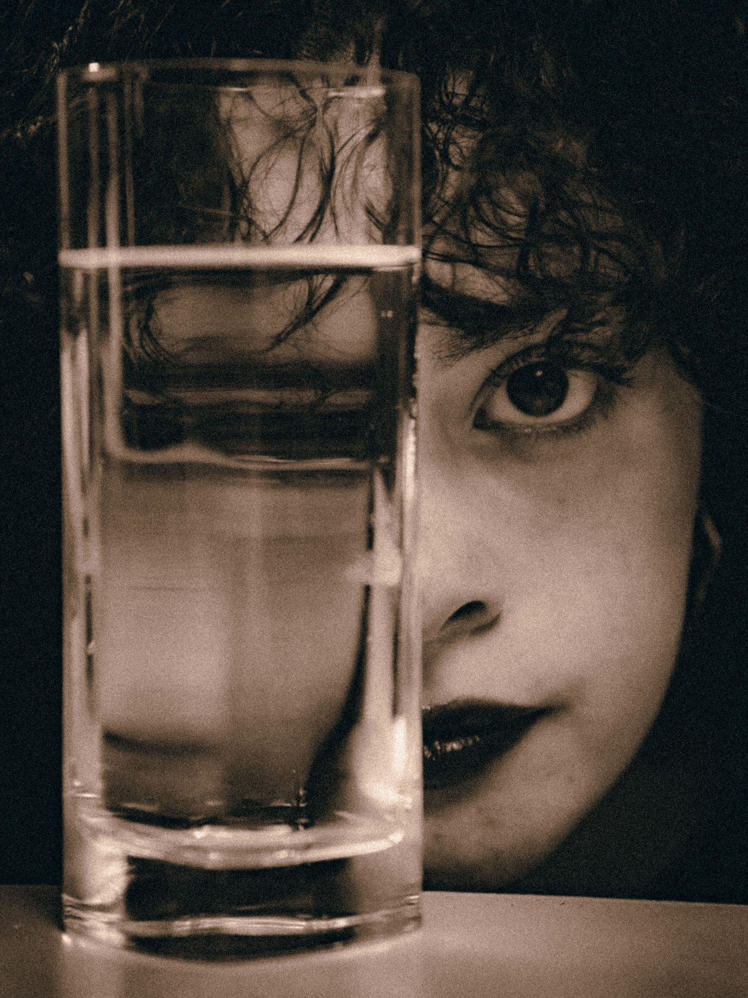 Close-up of a woman's face partially obscured behind a tall glass of water in moody sepia.