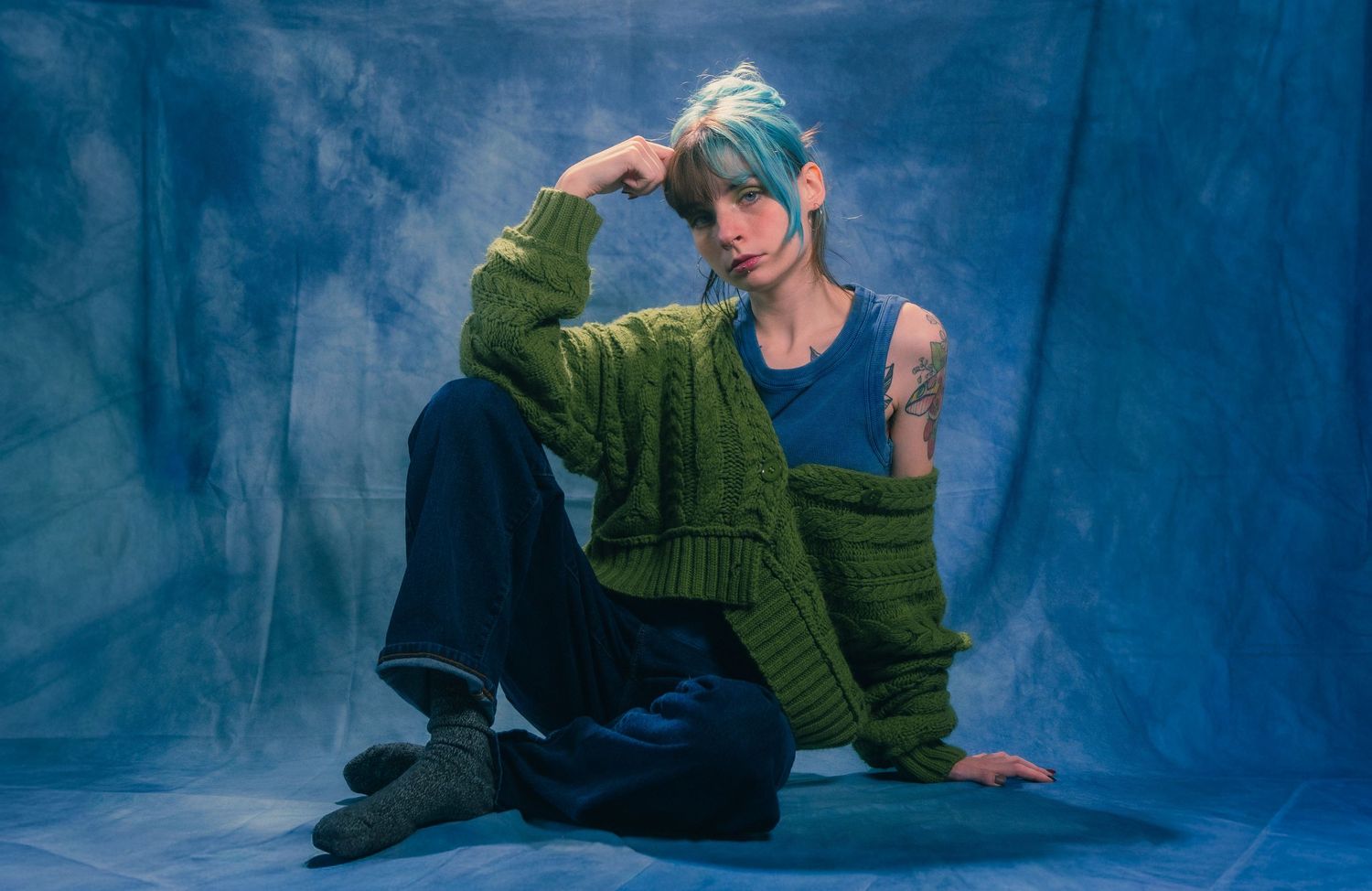 Model with blue hair poses in olive green cardigan and navy pants against blue fabric backdrop.
