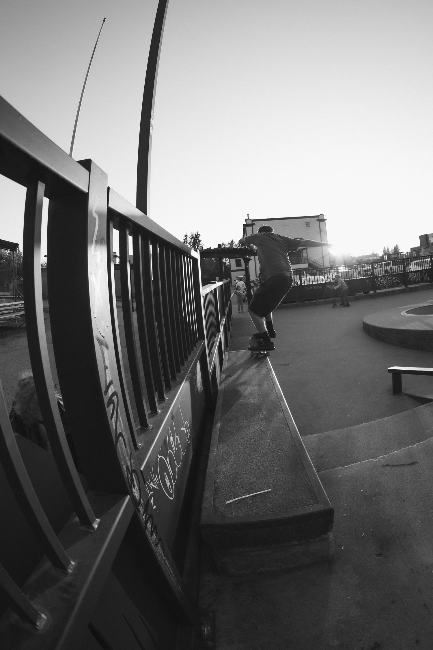 Black and white photo of a skateboarder jumping over a railing at a skate park during sunset.
