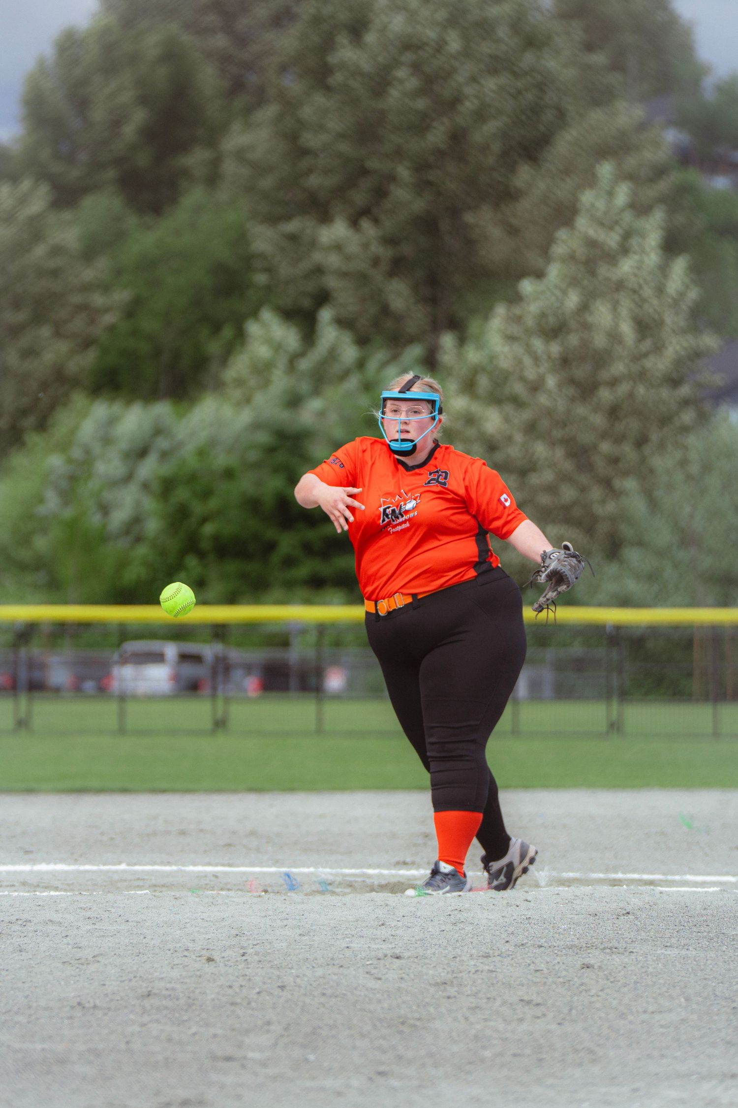 Female softball pitcher in orange uniform releases yellow ball toward batter on cloudy game day.