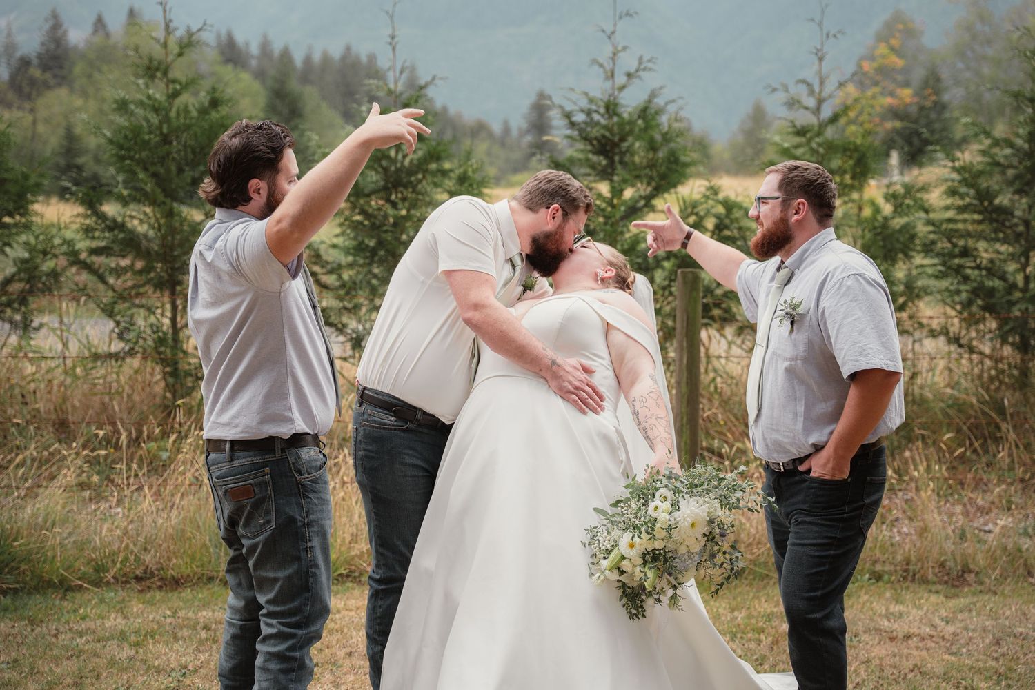 Bride and groom share a kiss while two groomsmen toast them outdoors at a rustic mountain wedding.