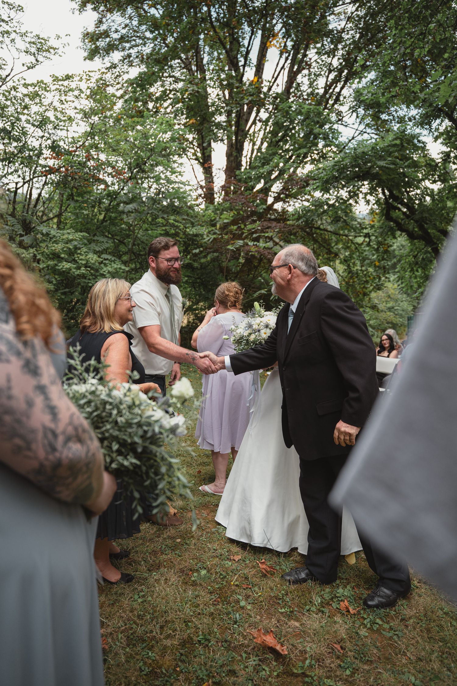 Groom in white shirt shakes hands with older man in black suit at outdoor woodland wedding ceremony.