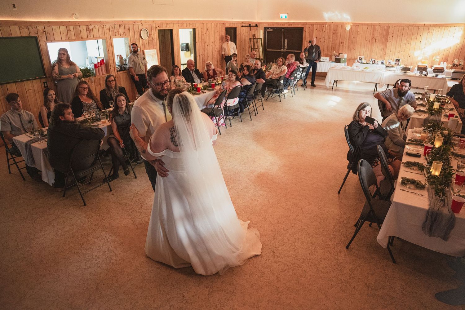 Bride and groom share their first dance at a rustic wedding reception with guests seated at decorated tables.