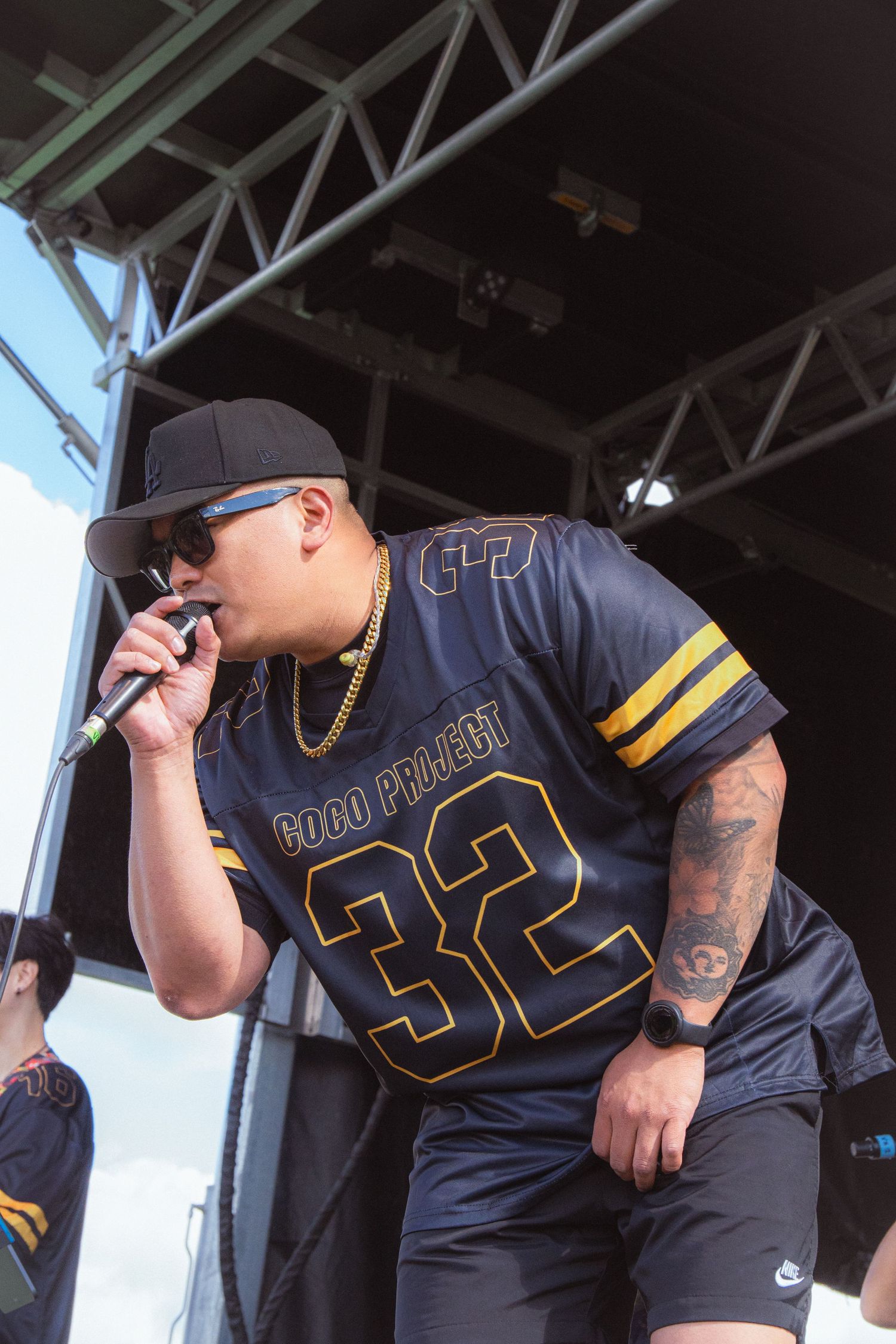 Male rapper in Coco Project number 32 jersey and cap performs with microphone at outdoor festival.
