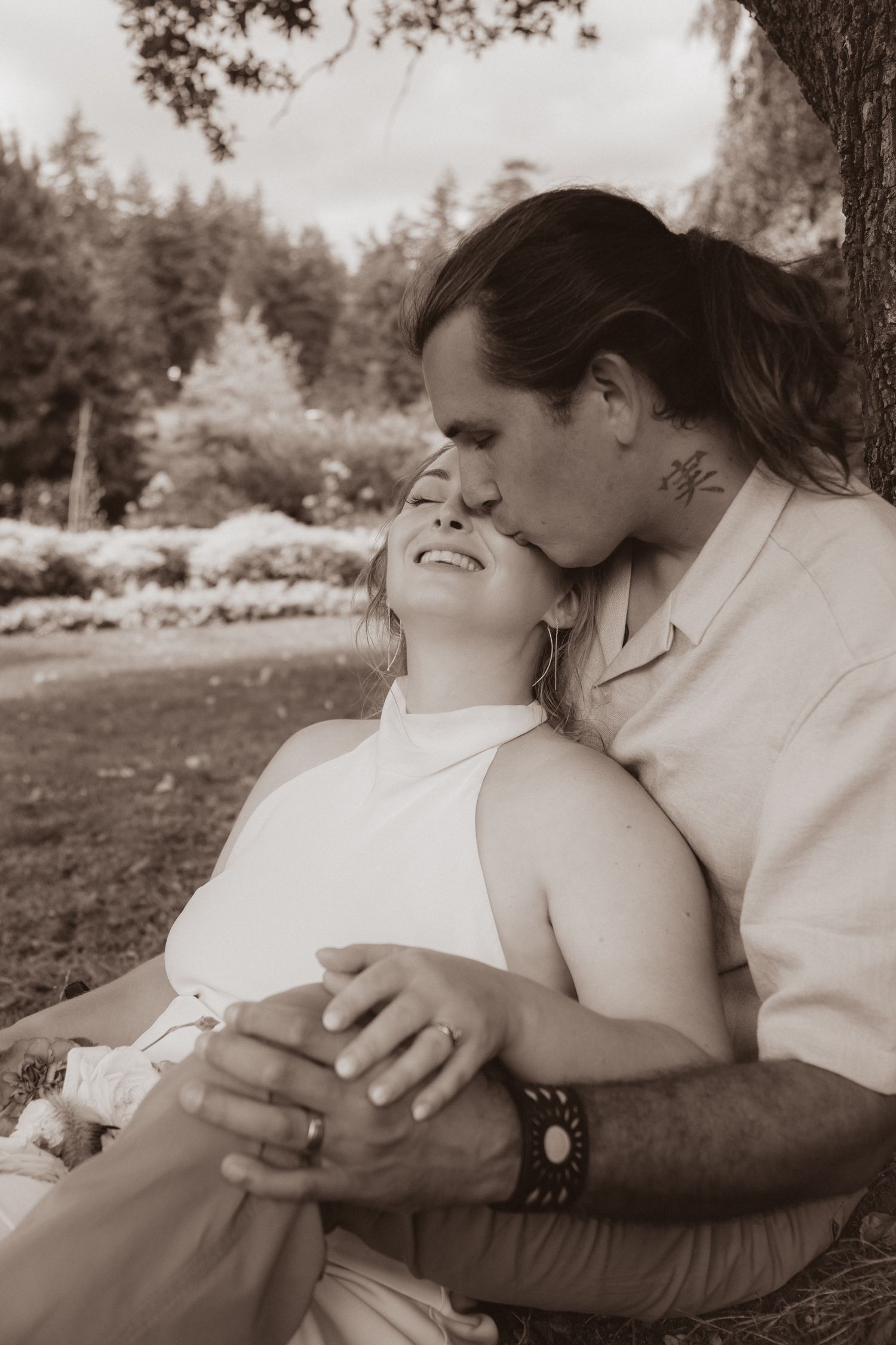 Sepia-toned photo of a couple embracing under a tree, man kissing woman's cheek as she smiles in a garden.