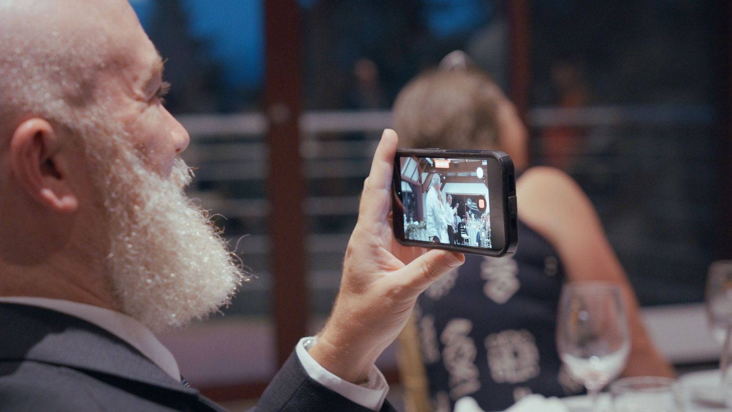 Elderly man with white beard photographing wedding reception on smartphone.
