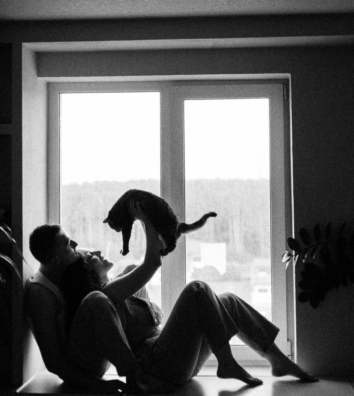 Black and white photo of couple sitting by window lifting cat in air, cozy home interior with natural light.