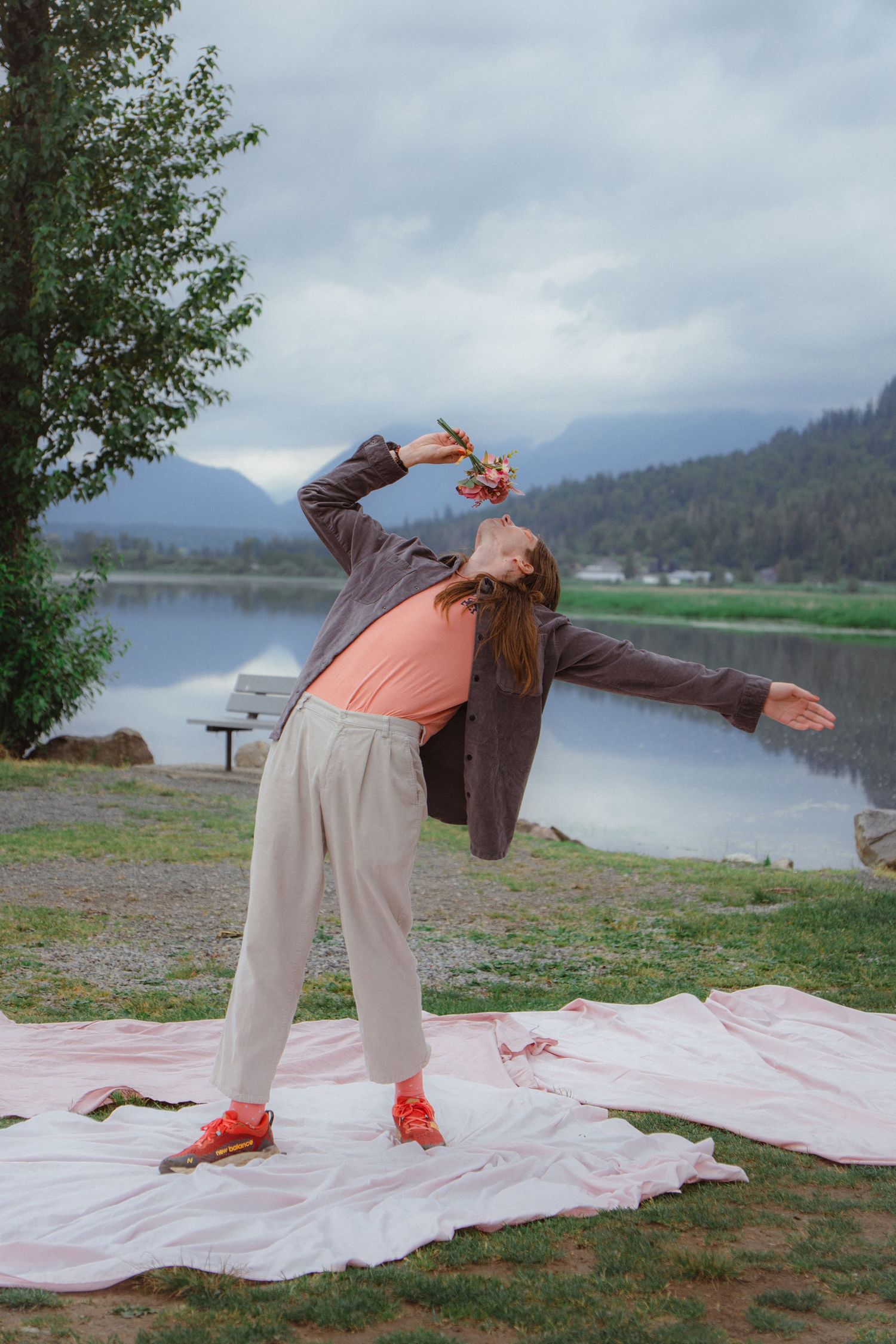 Woman in gray jacket and beige pants poses dramatically by a mountain lake on a pink blanket.