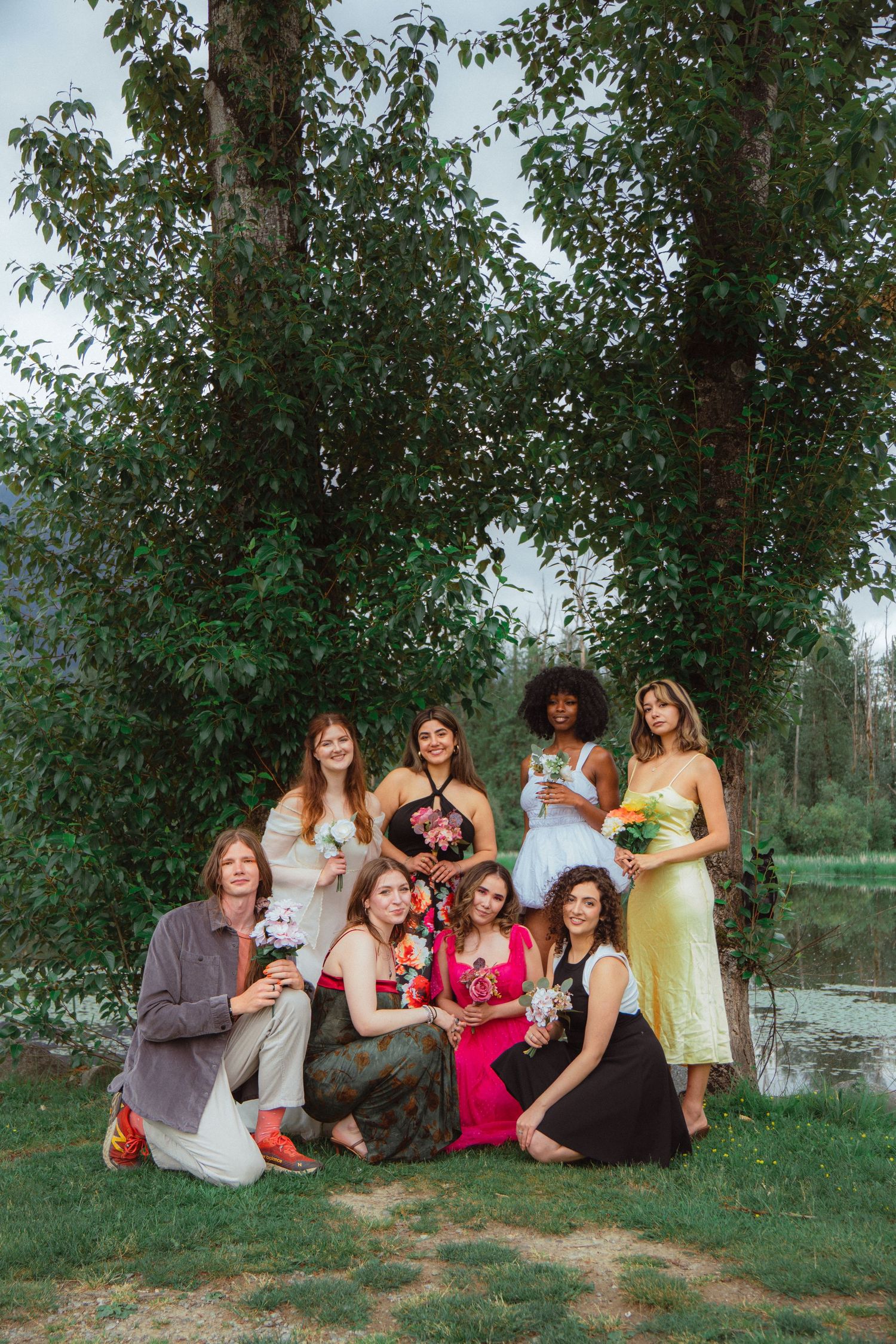 Group of eight diverse women in colorful dresses posing with bouquets near lakeside trees.
