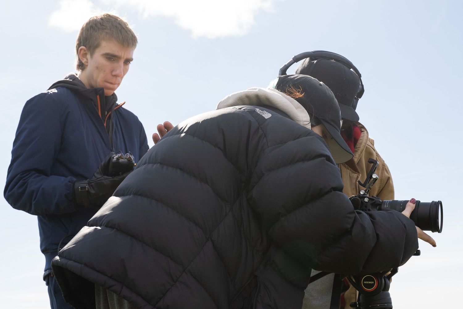 Young man in a light hoodie reviewing content with a camera operator outdoors under a blue sky.