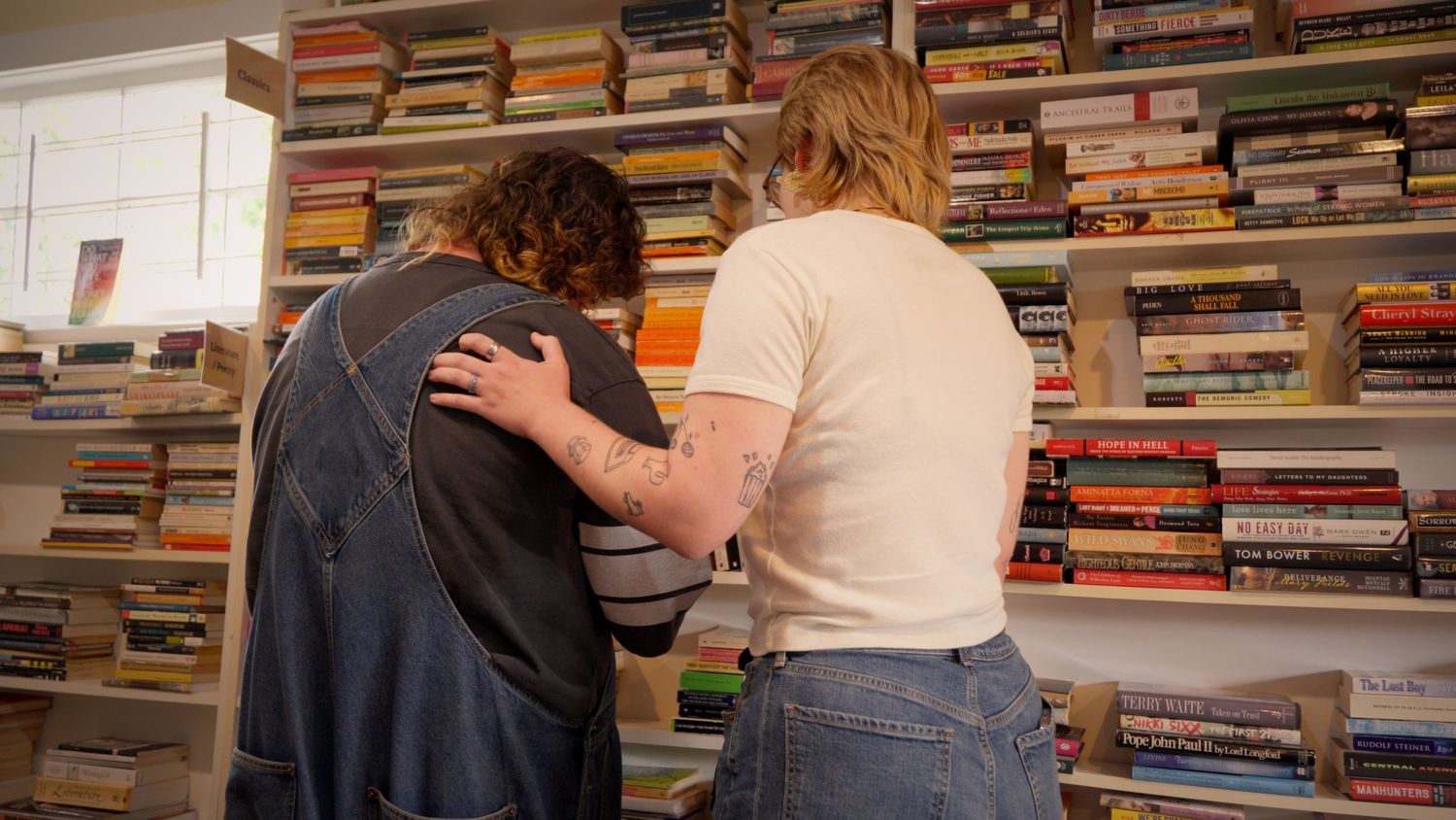 Two people standing close together browsing a large wall of colorful stacked books.