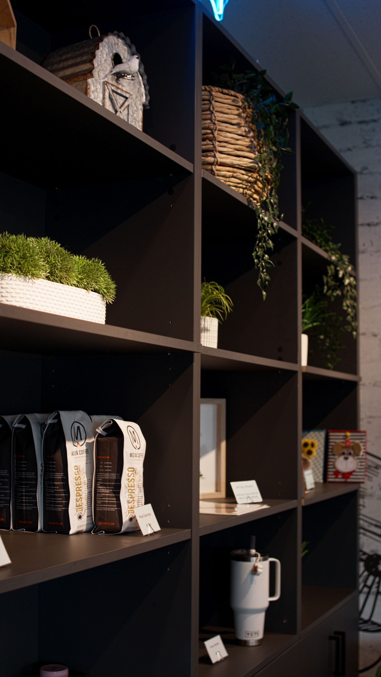 Dark retail shelving unit displaying espresso coffee bags, plants, tumblers, and decorative items.