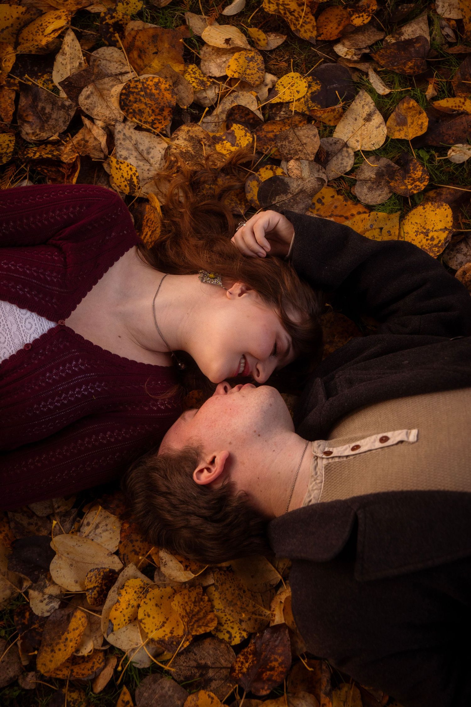 Romantic couple lying in colorful fall leaves, smiling and leaning close together in a tender moment.
