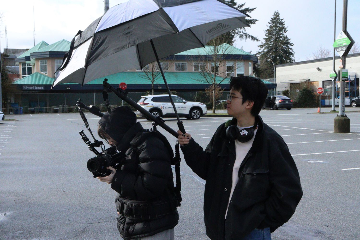 Two film crew members work outdoors, one holding umbrella over camera in overcast weather.