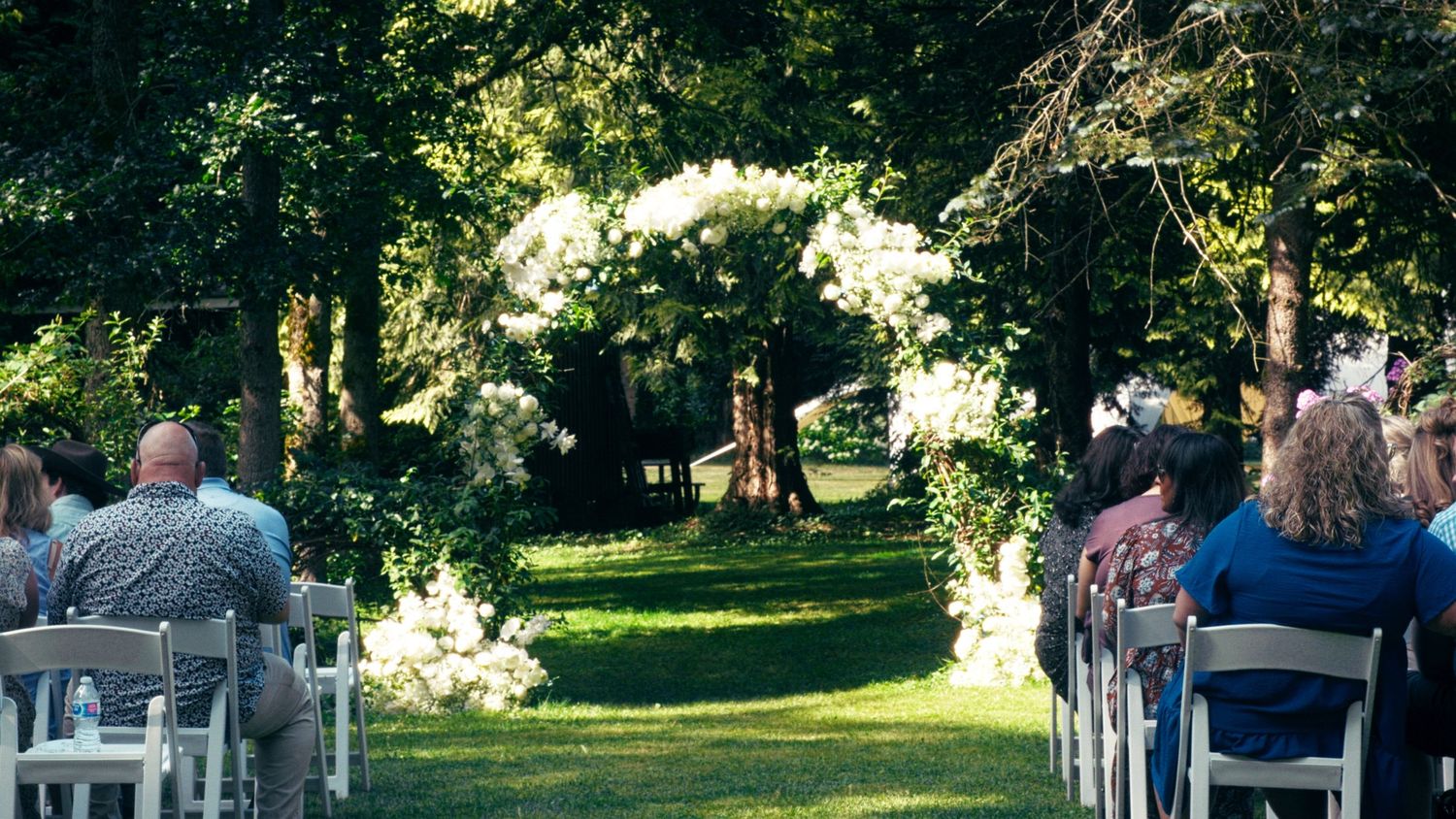 Outdoor wedding ceremony with white floral arch, guests seated in white chairs on green lawn surrounded by trees.