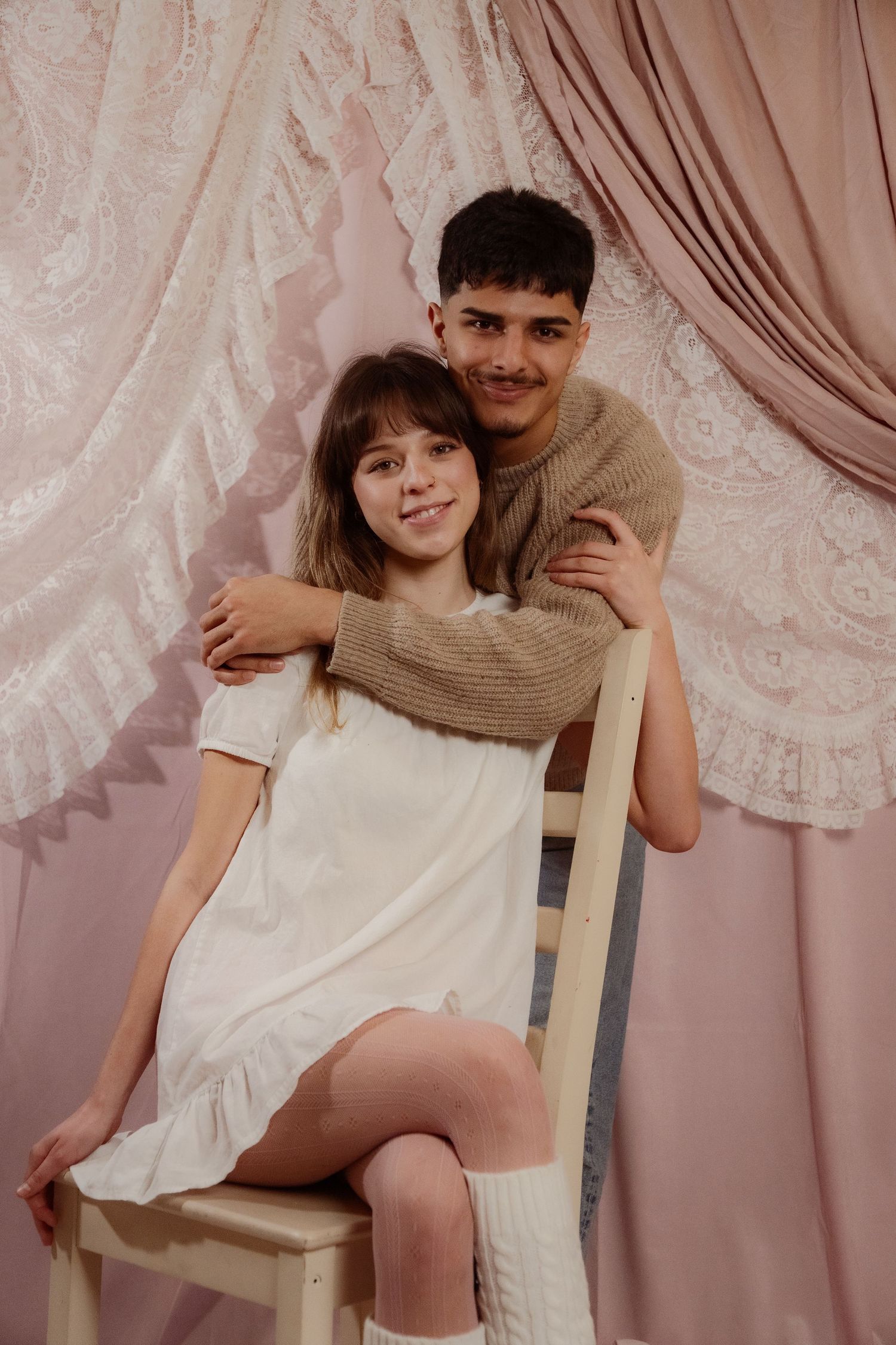 Young couple posing together, man in knit sweater hugging woman in white dress on a chair.