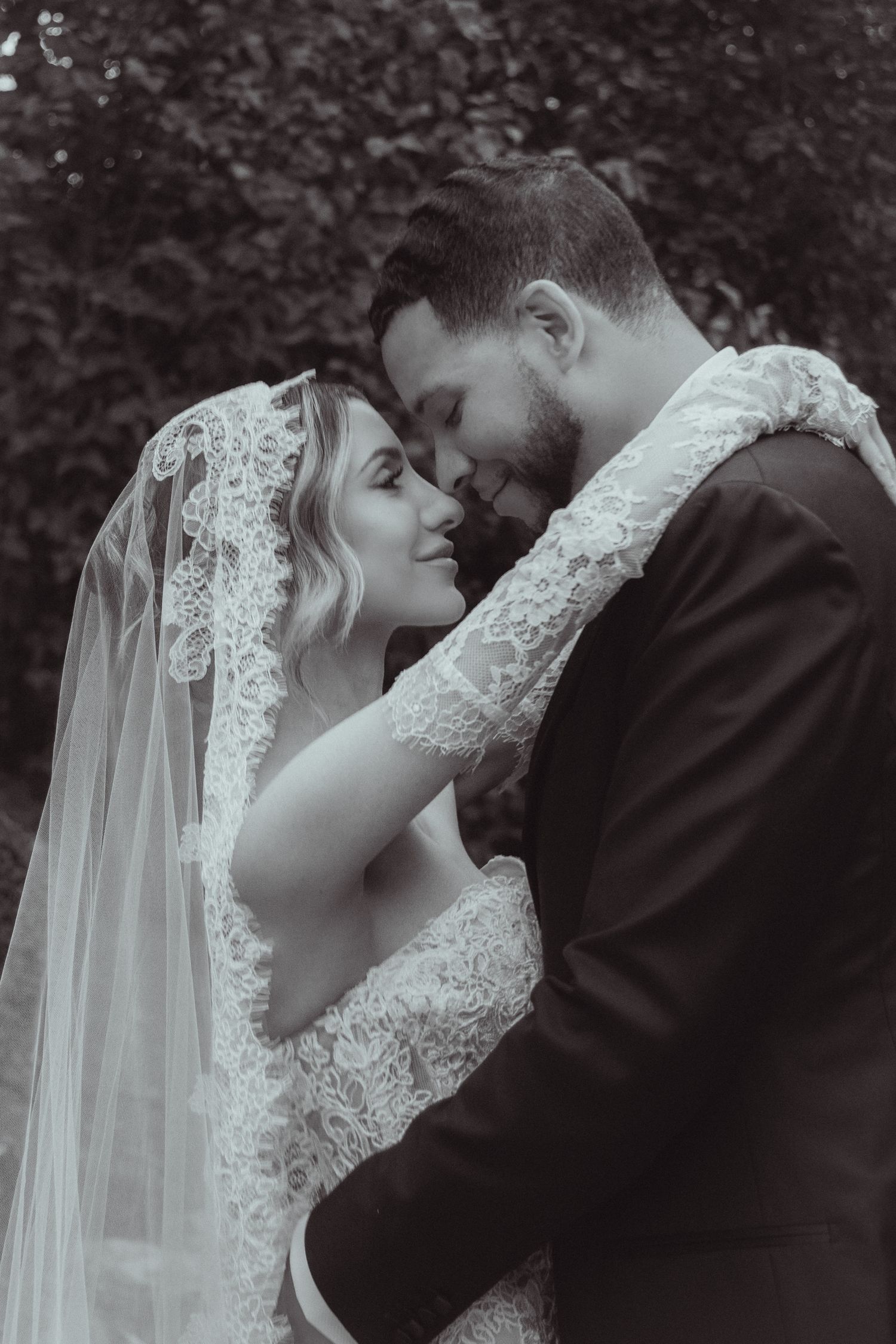 Black and white photo of a bride and groom sharing an intimate moment, her lace veil draped over his shoulder.