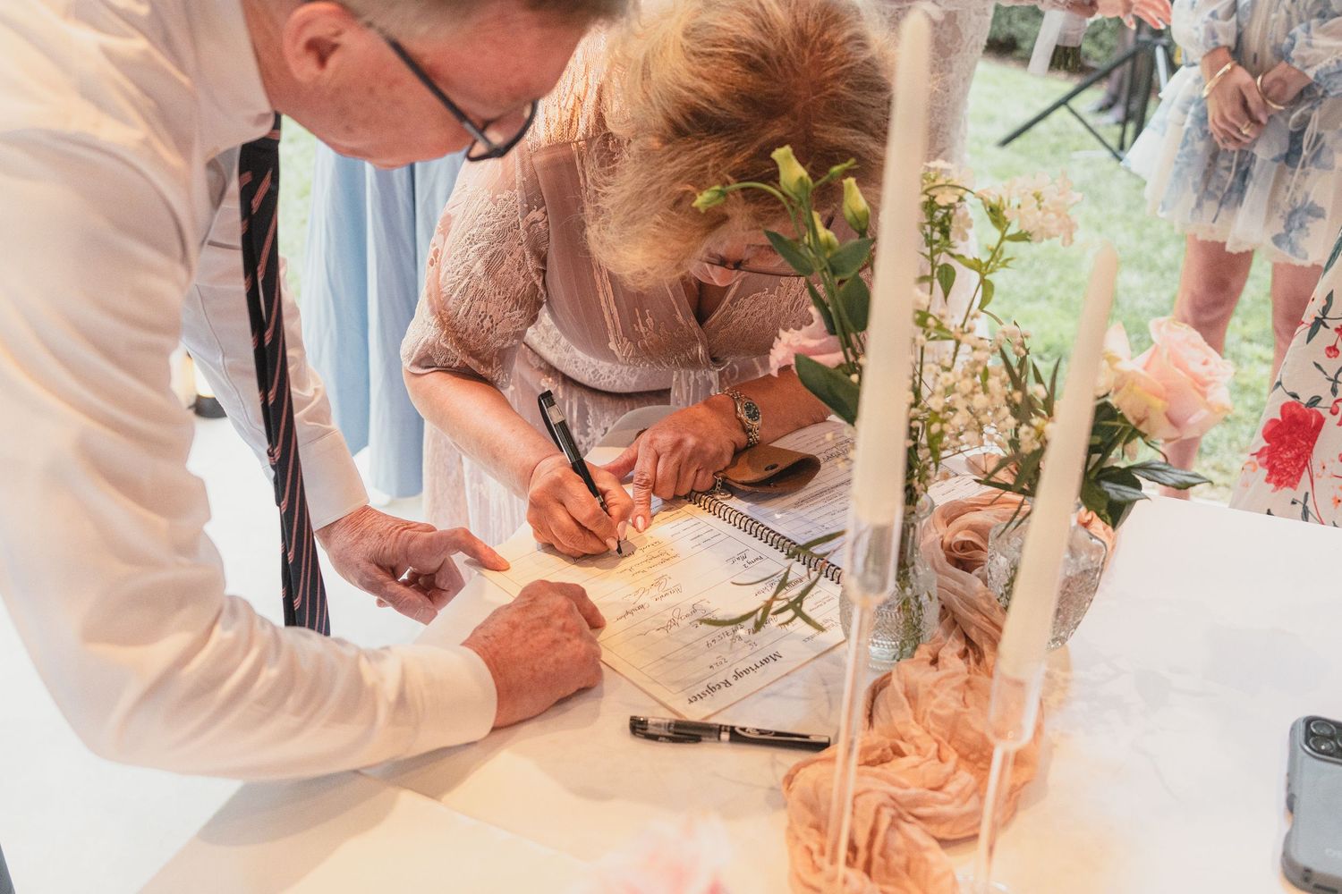 Wedding guests signing the guest book at a decorated reception table with floral arrangements and candles.