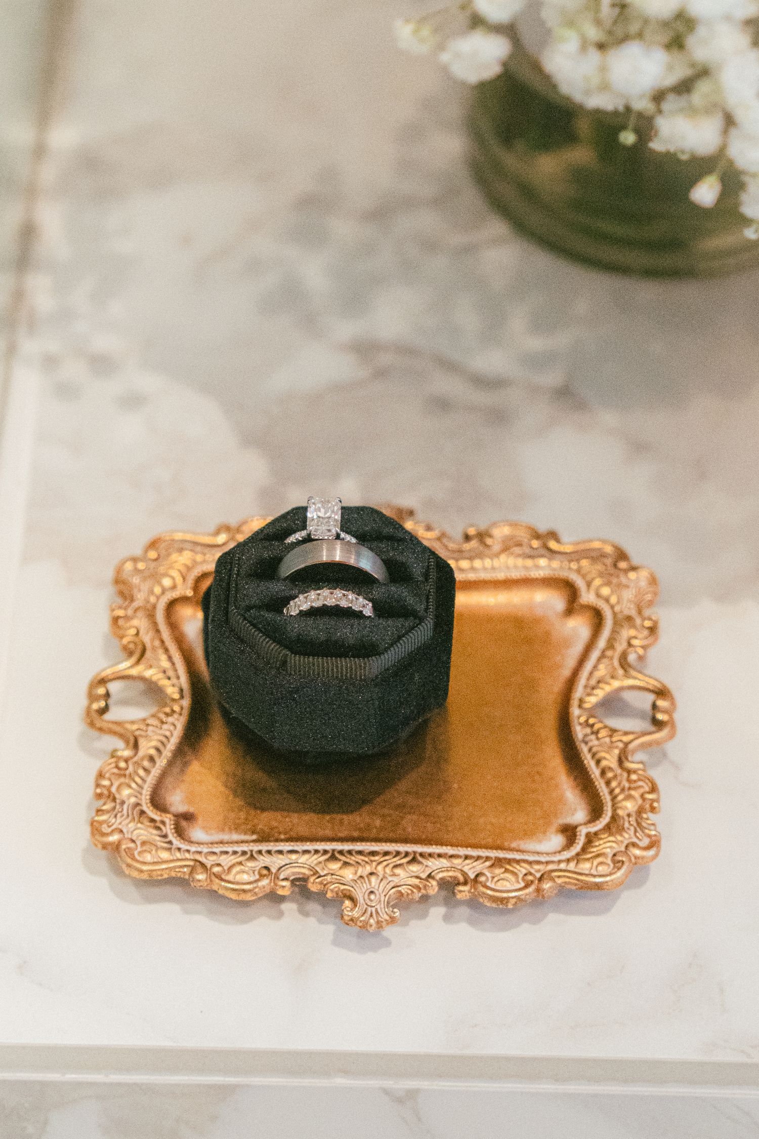Two diamond wedding rings displayed in a dark green velvet box on an ornate gold tray atop a marble surface.