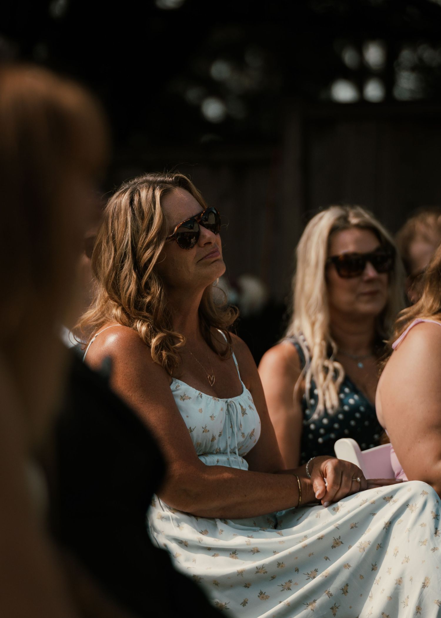 Women in sunglasses and summer dresses seated outdoors at a sunny gathering or event.