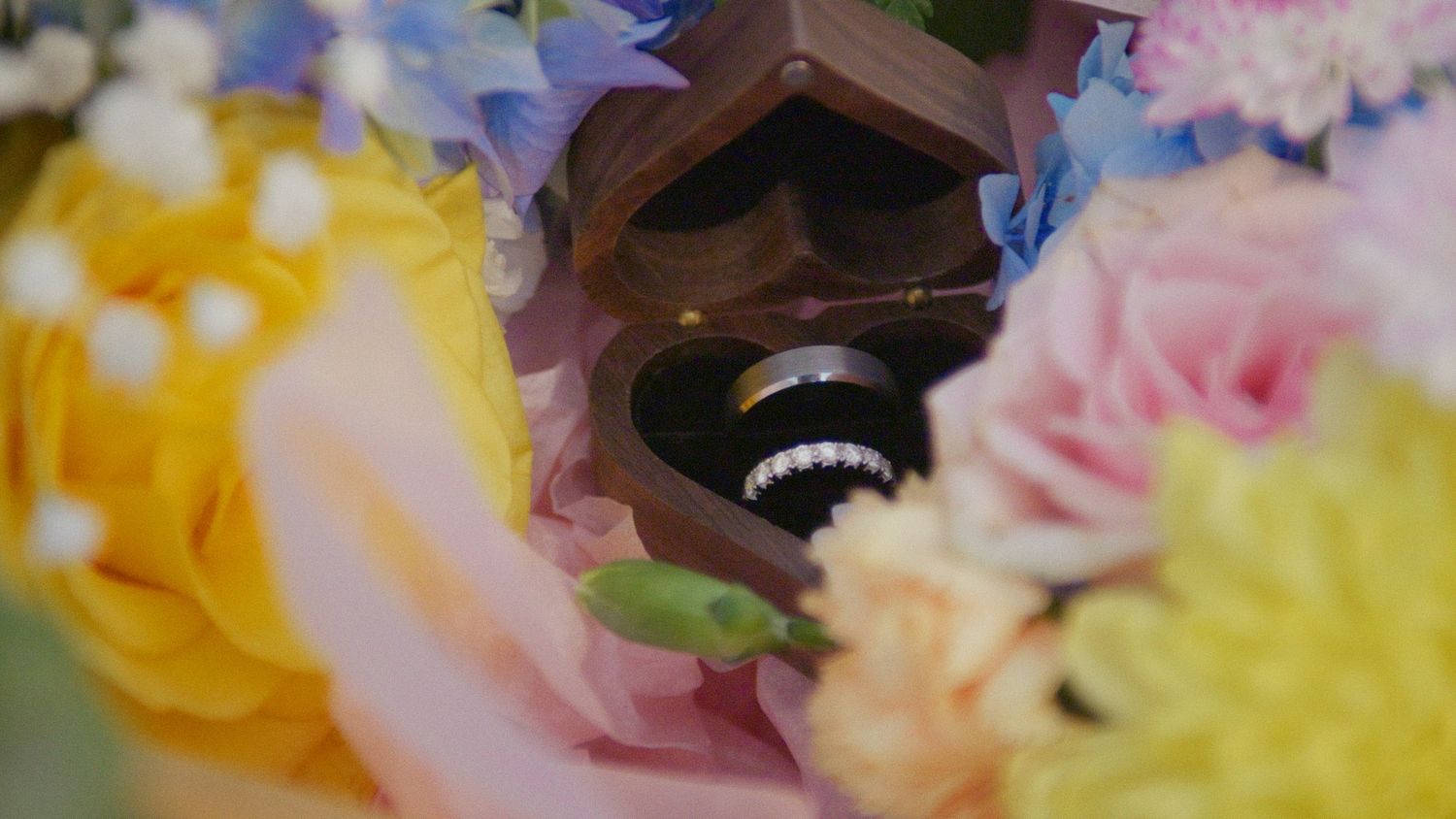 Close-up of wedding rings in a wooden box surrounded by colorful pink, yellow, and blue flowers.