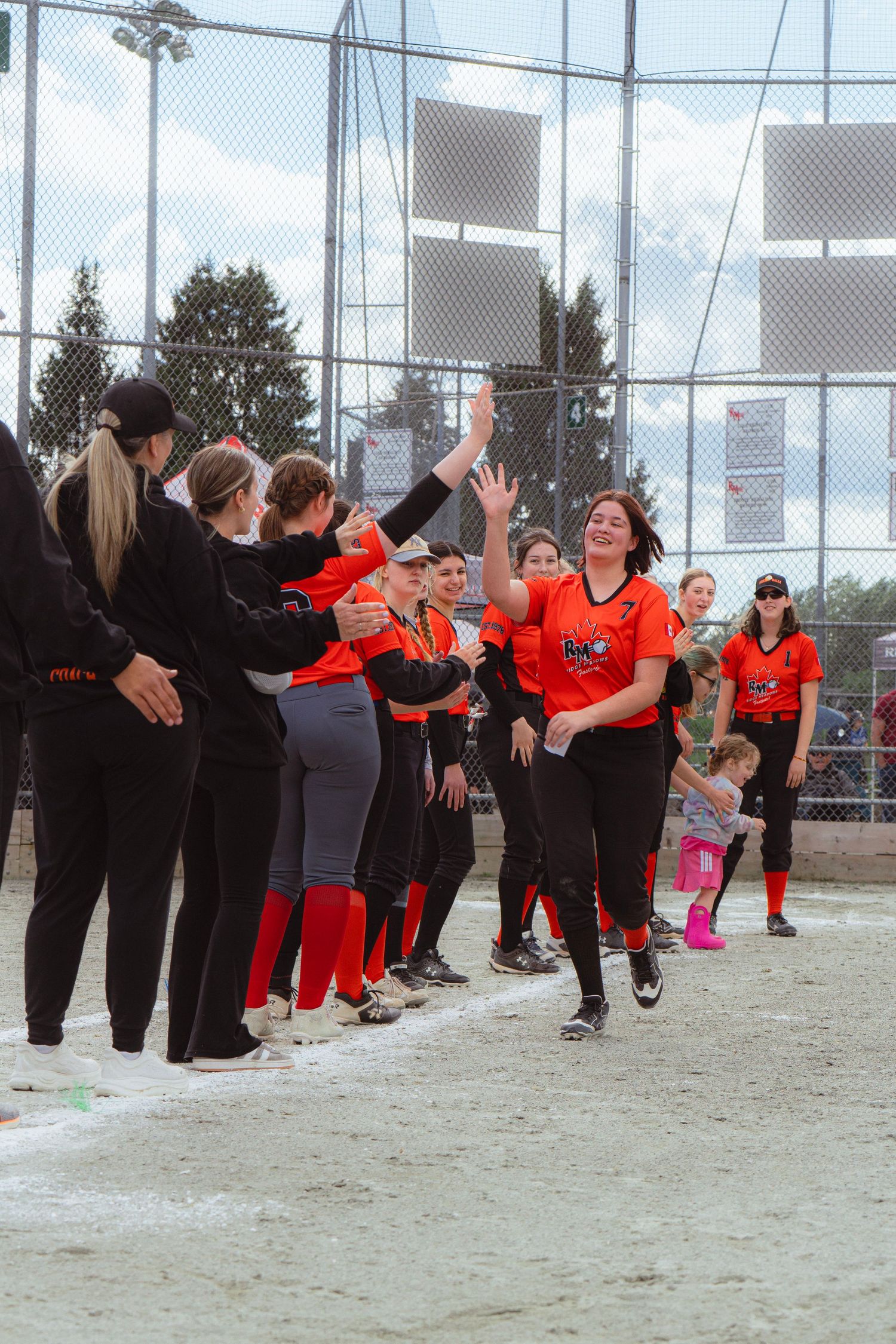 Smiling softball player in orange jersey high-fives teammates lined up along the dugout after scoring.