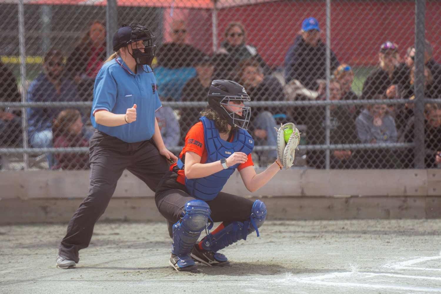 Softball catcher in blue protective gear crouches at home plate beside umpire during outdoor game.