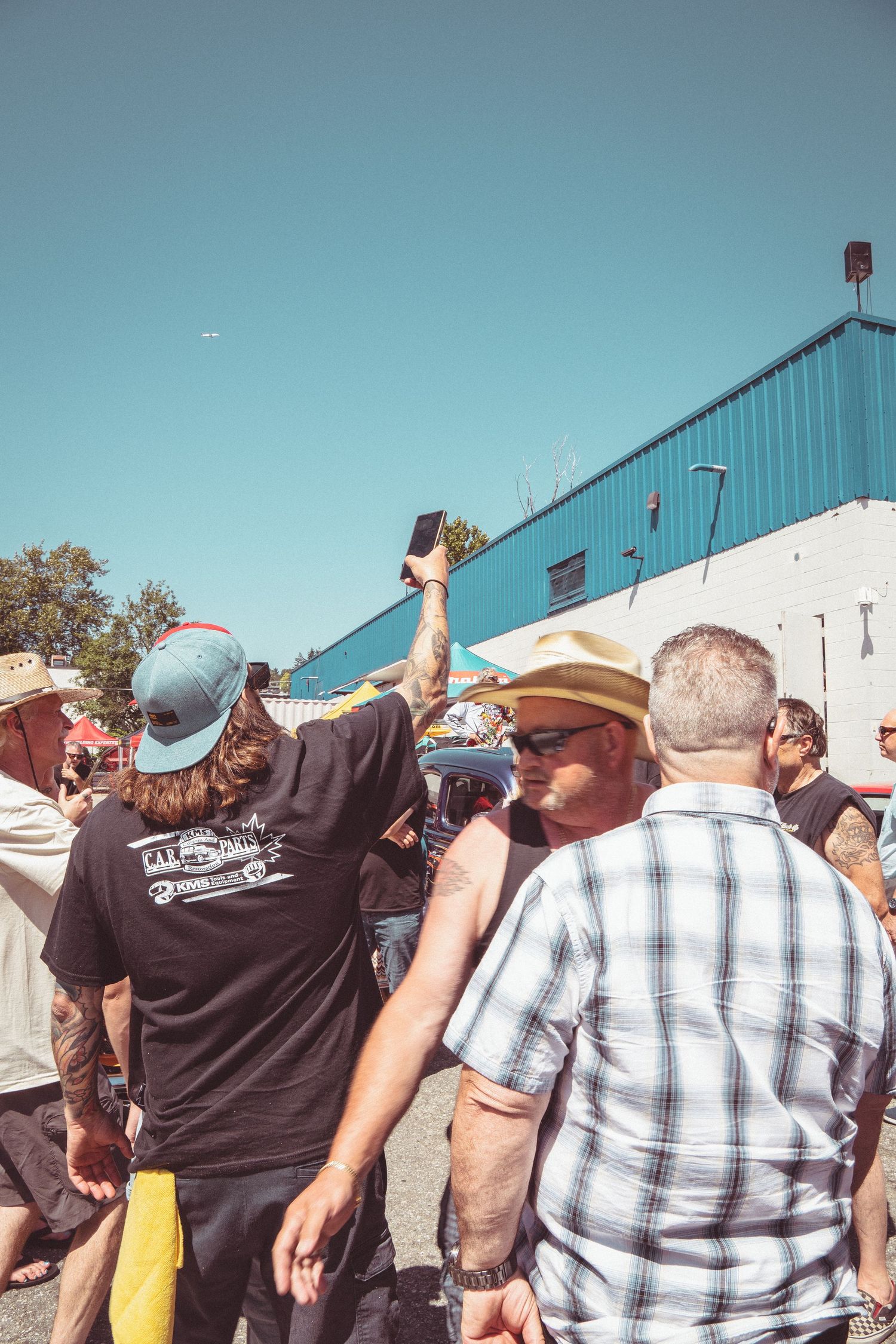 Crowd of men at outdoor car show, one raising phone to take photo on sunny day.