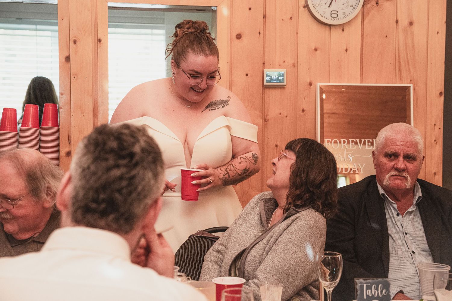 Tattooed bride in white off-shoulder gown mingles with seated guests holding a red cup at wedding reception.