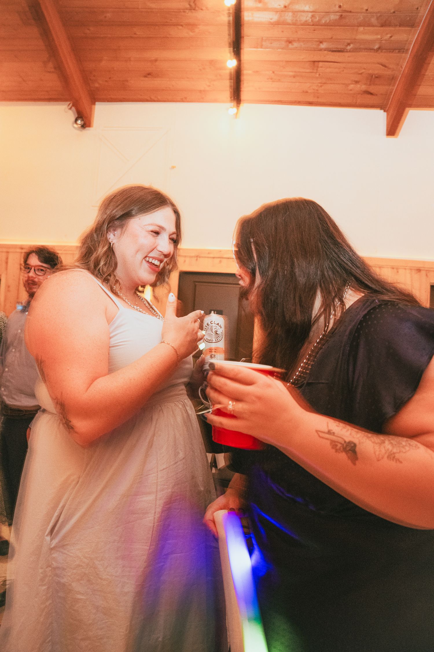 Two women laughing and holding drinks at an indoor party with colorful lights and wood-paneled ceiling.