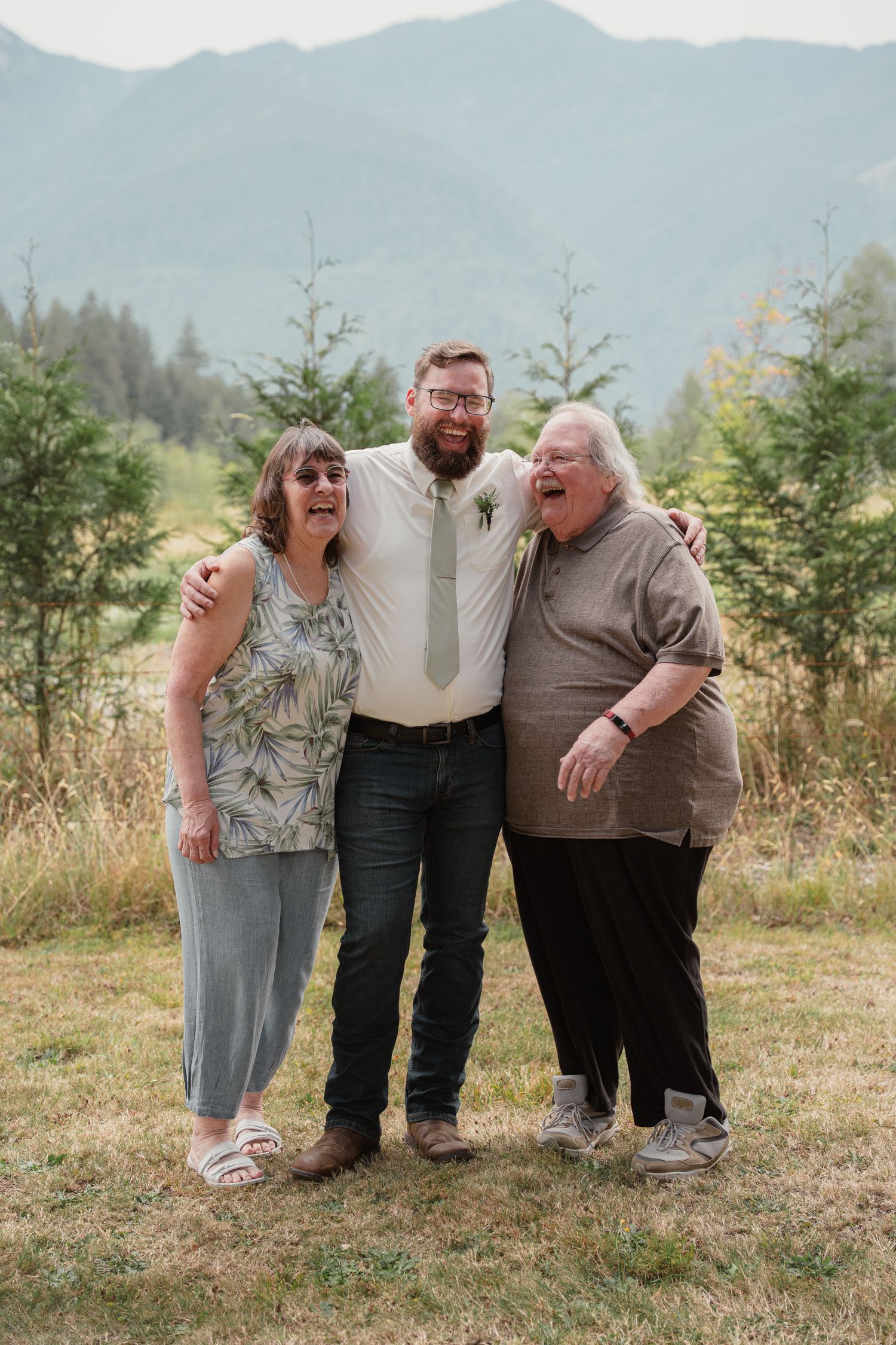 Groom with boutonniere hugs two older women outdoors at mountain wedding venue.
