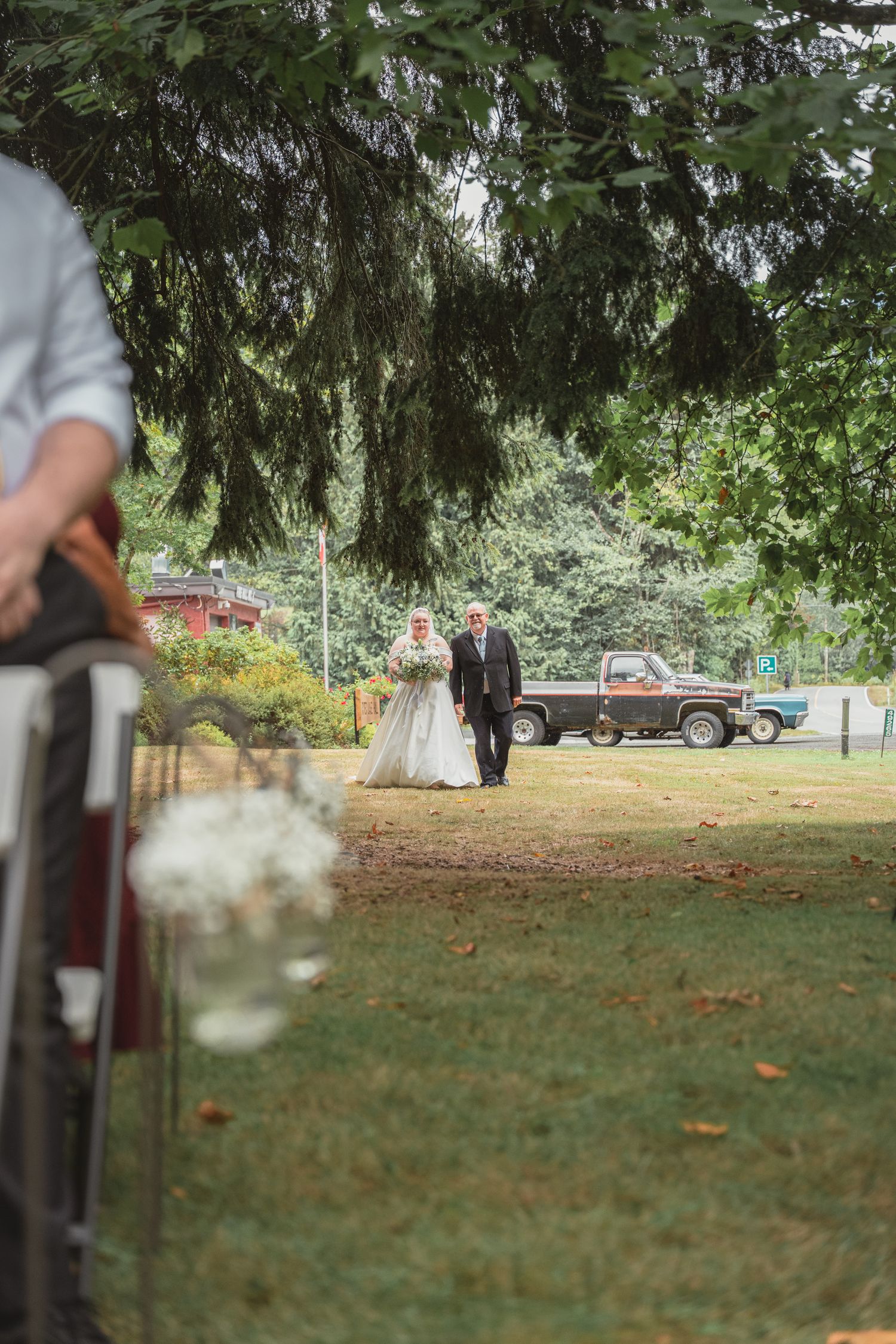 Bride walking down outdoor aisle with escort toward rustic woodland wedding ceremony on green lawn.