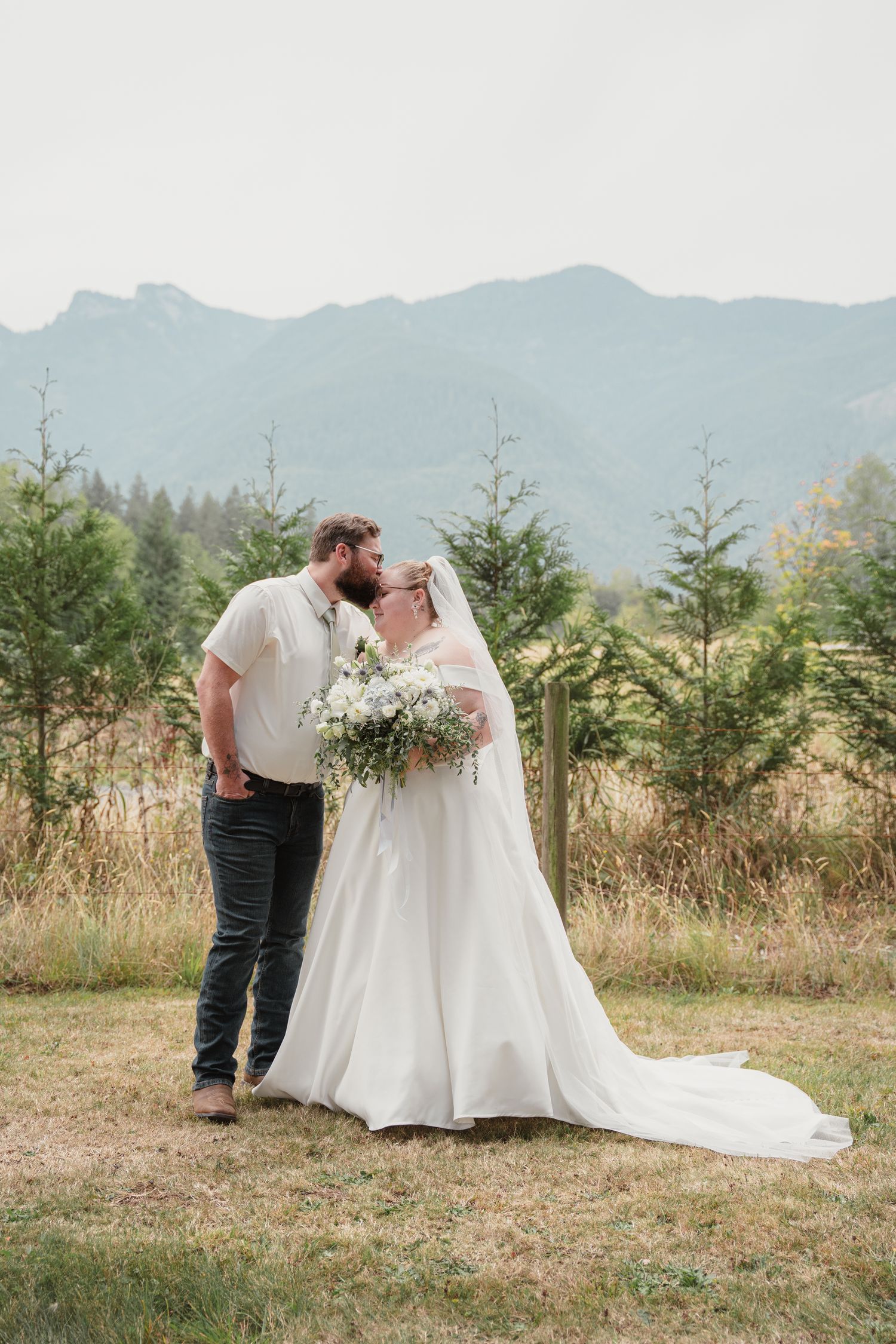 Bride and groom share a kiss outdoors with mountain views, evergreen trees, and golden fields in the background.