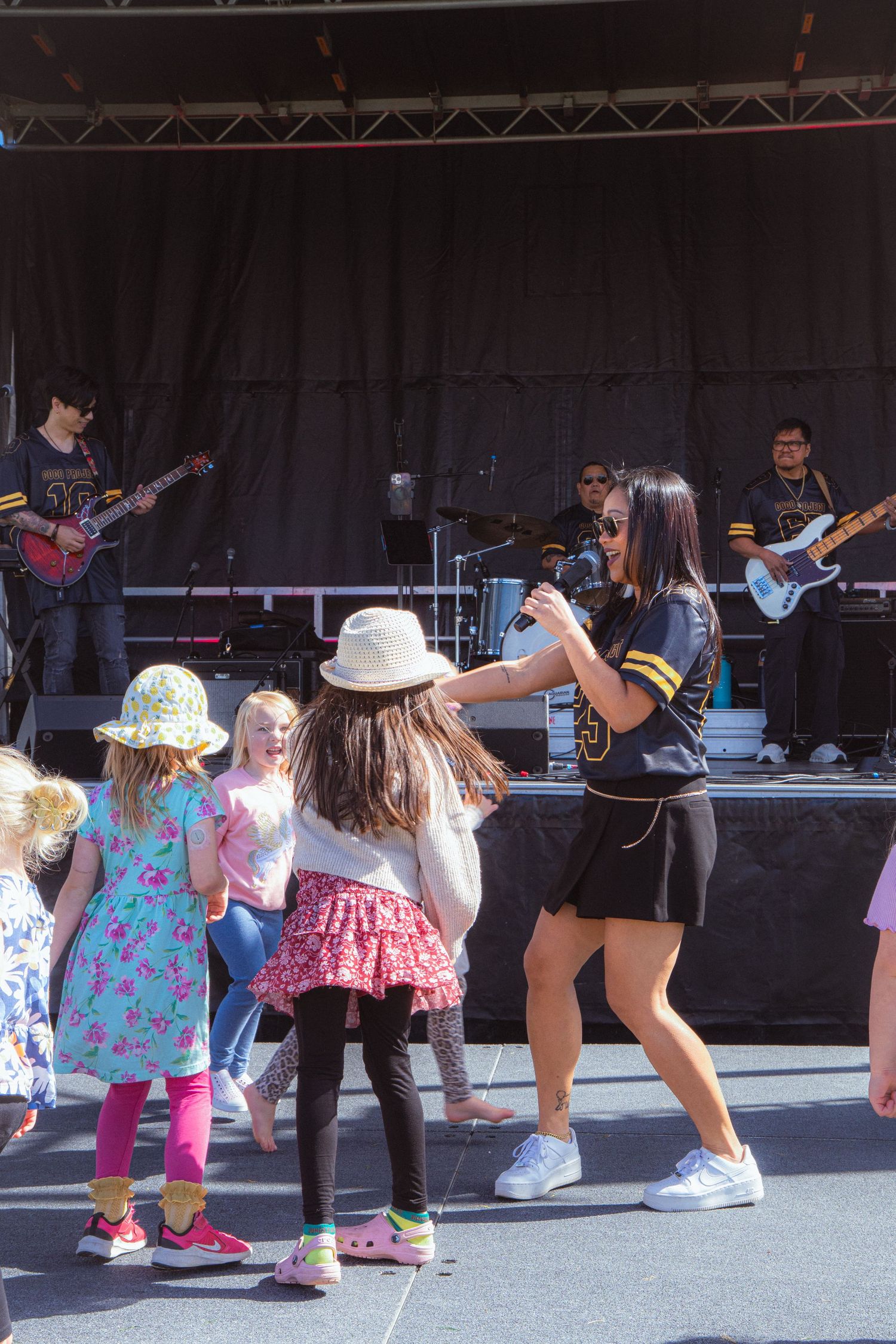 Energetic female performer engages with kids dancing near an outdoor concert stage with live band.