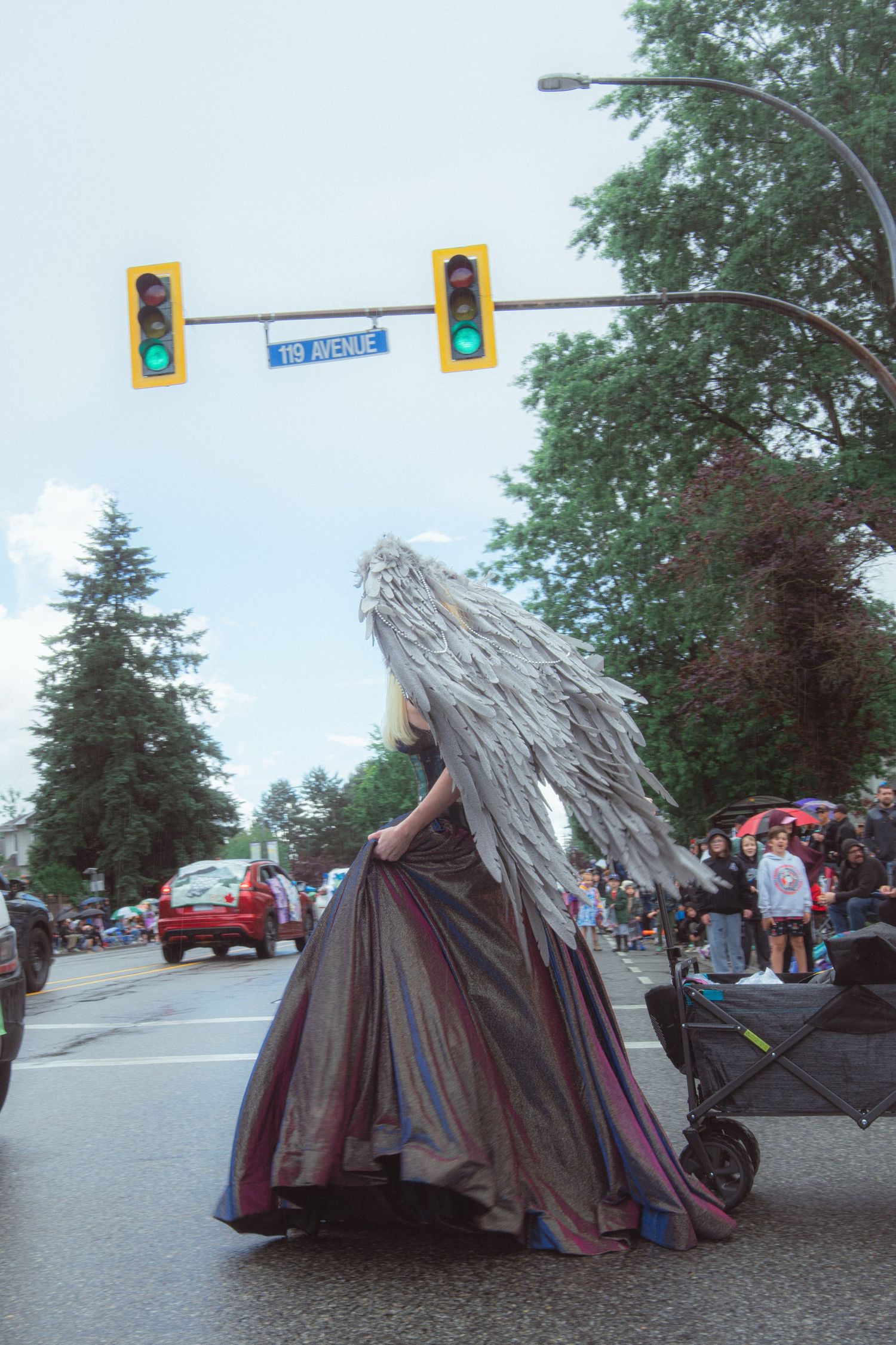 A performer in a dramatic angel wing costume and long gown walks in a street parade.