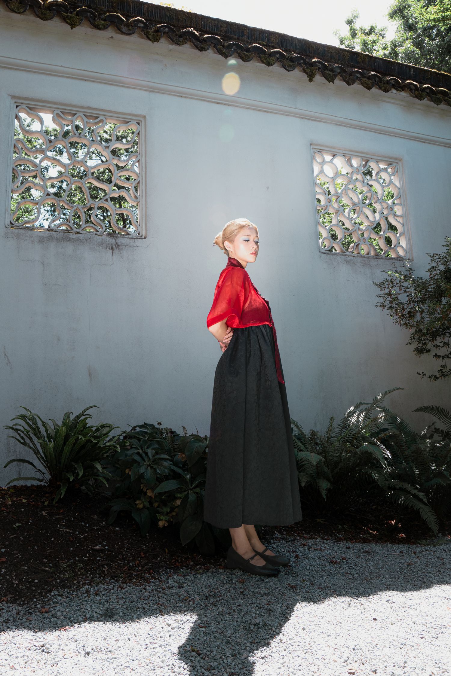 Woman in red blouse and dark maxi skirt posing by white wall with decorative windows.