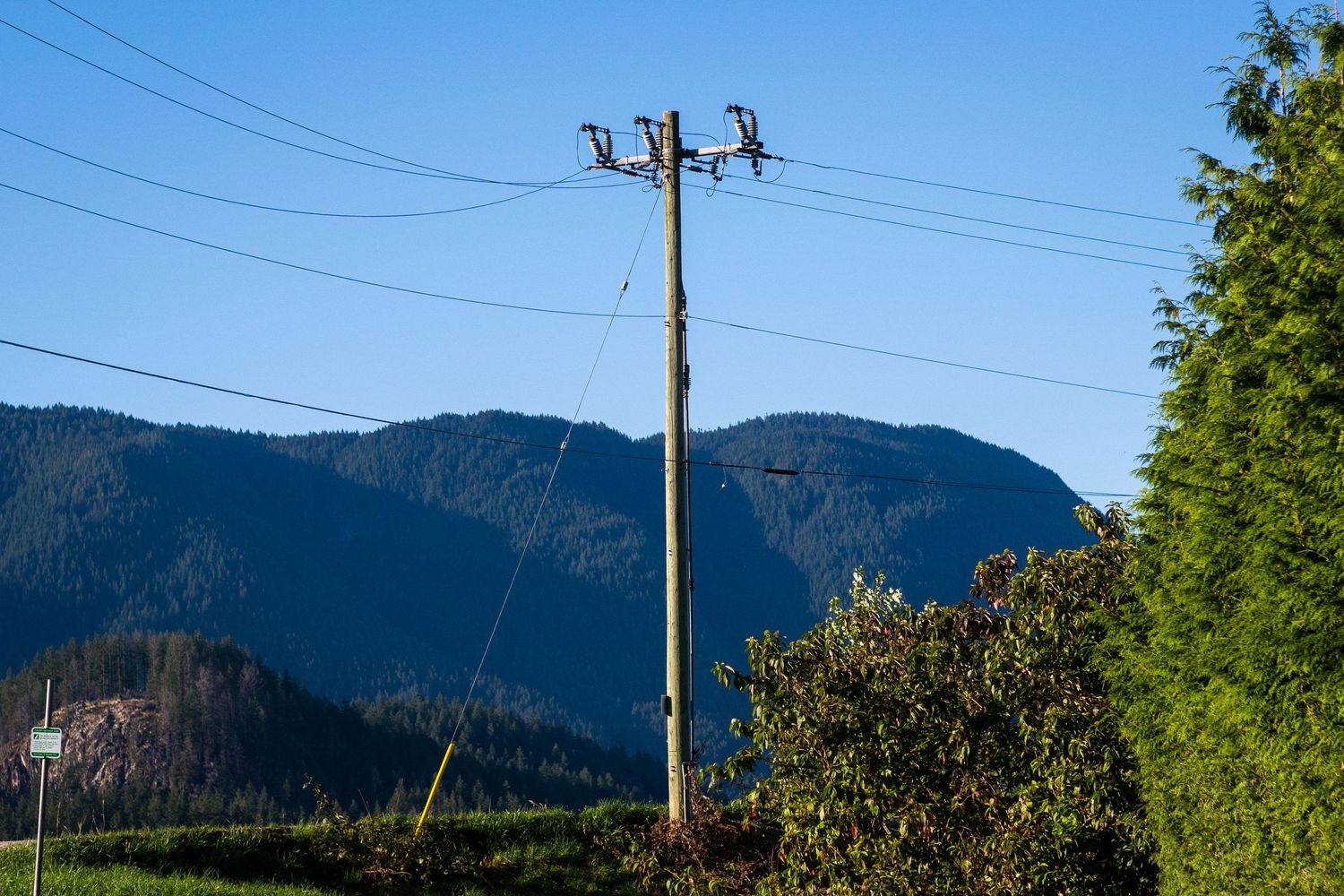 Utility pole with power lines against a clear blue sky with forested mountains in background.